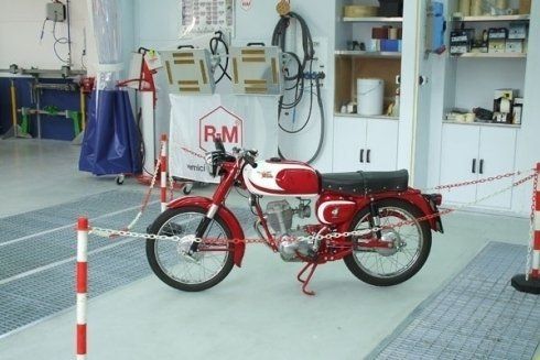 Motocicletta d'epoca rossa esposta dietro transenne di corda in un ambiente museale o officina.