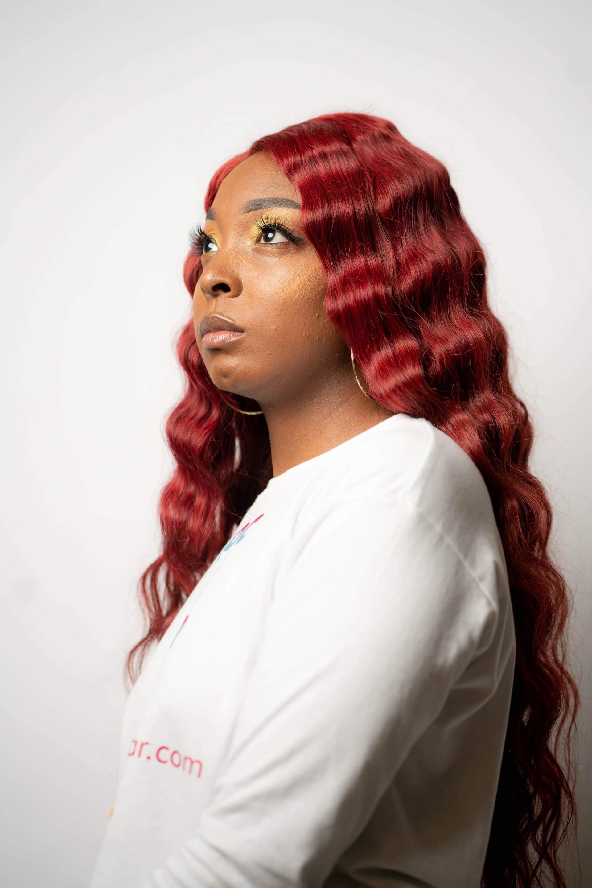 Side-profile portrait of a person with long wavy red hair wearing a white top against a light background