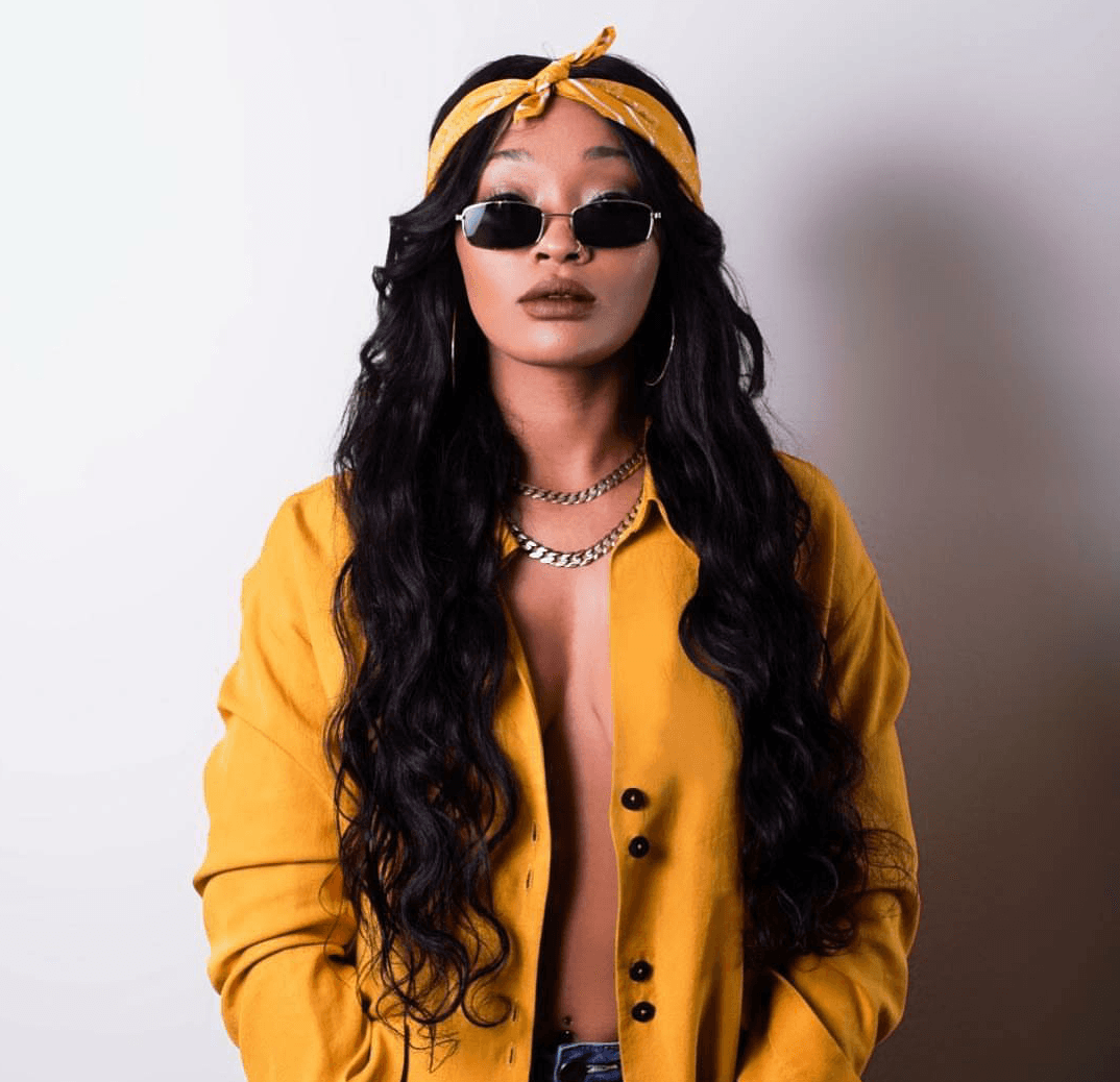 Woman in a yellow outfit and headscarf, wearing sunglasses against a plain light background