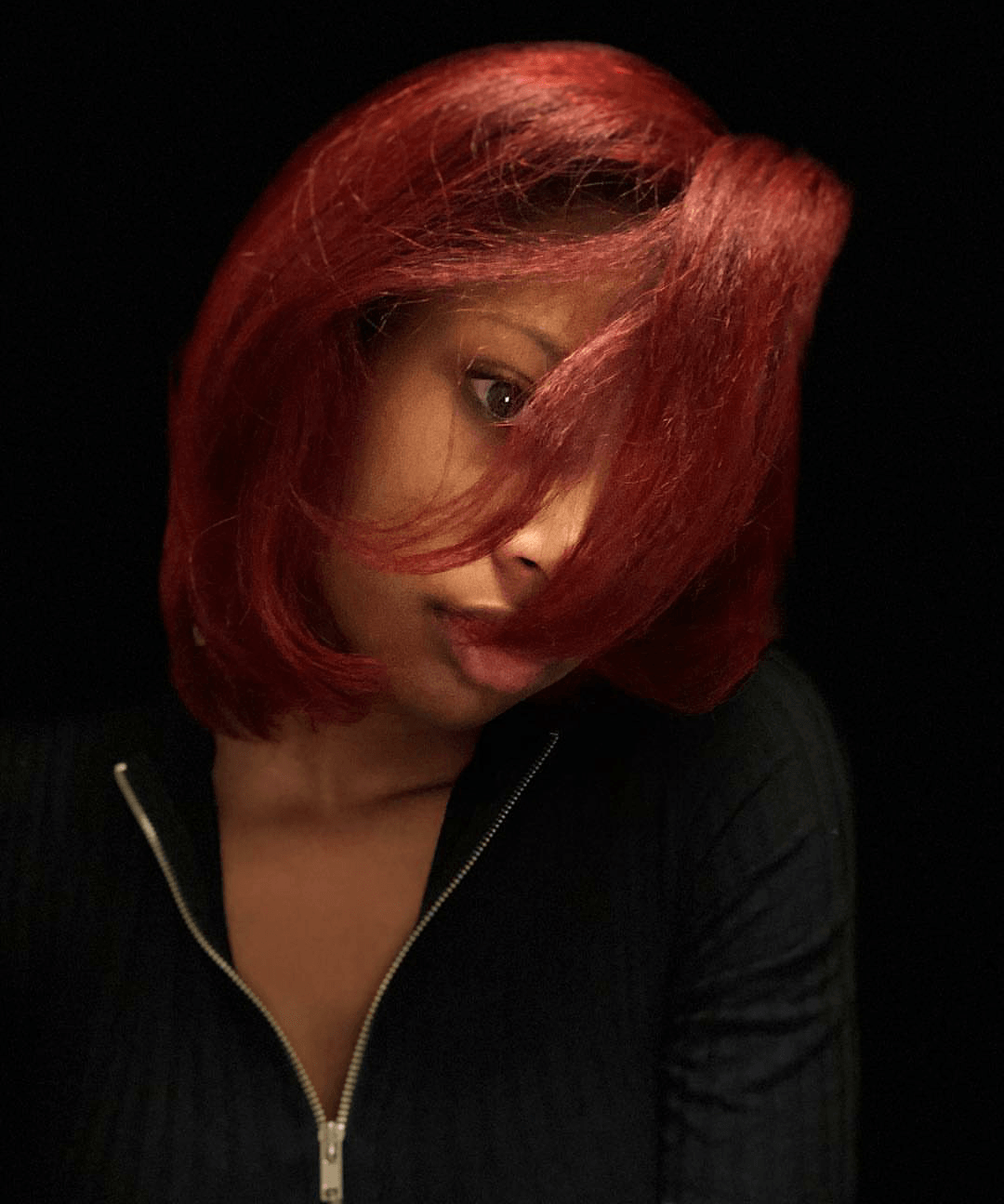 Portrait of a person with short red hair in a black zip-up top against a dark background