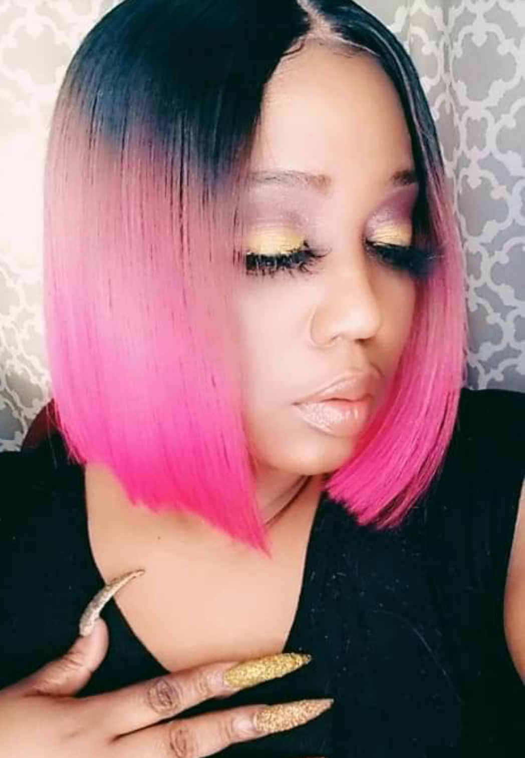 Woman with a pink ombré bob, gold eye makeup, and long gold nails, looking downward with hand on chest
