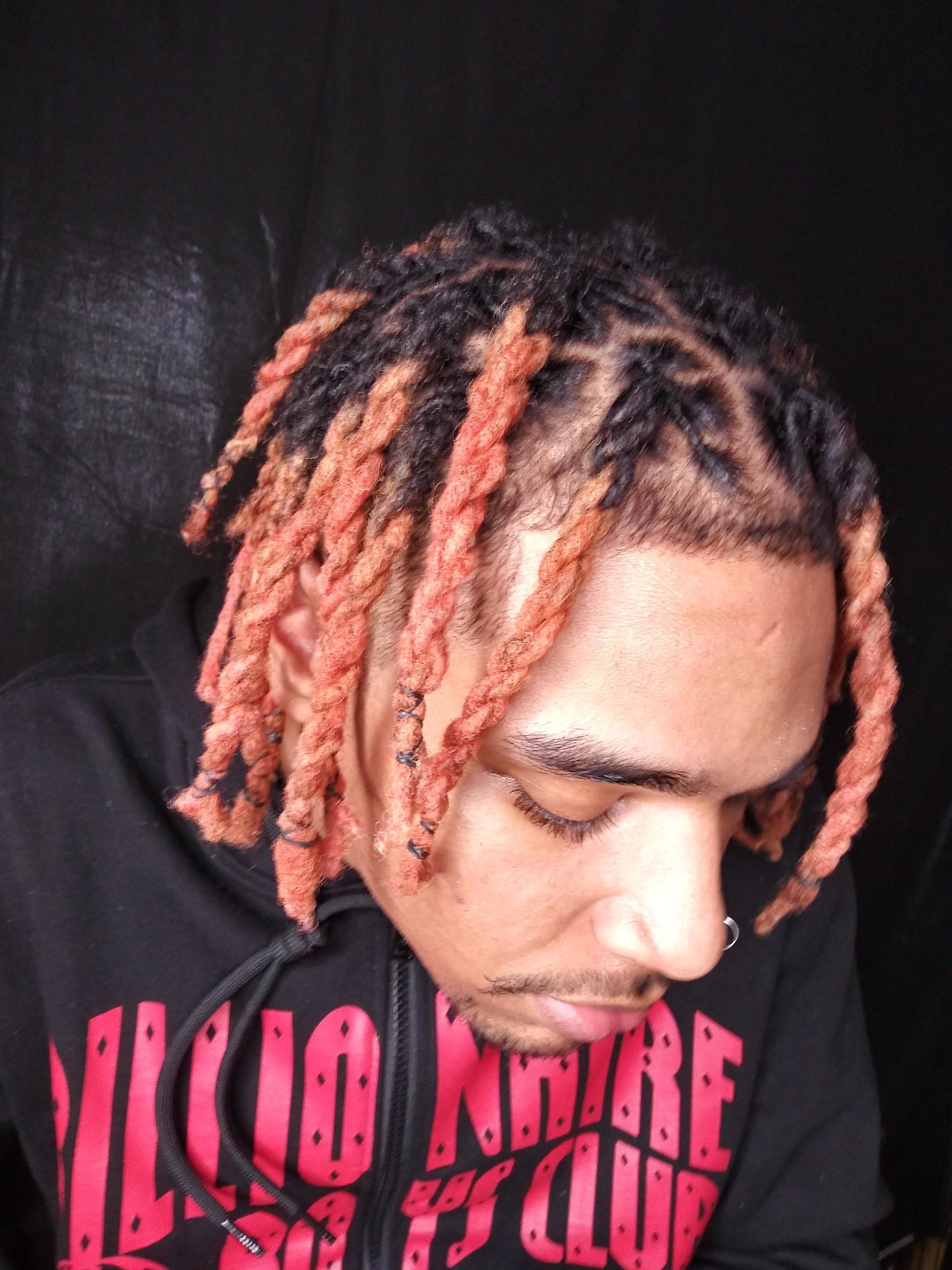 Person with pink dreadlocks looking down, wearing a black shirt with bright pink text, against a dark background.