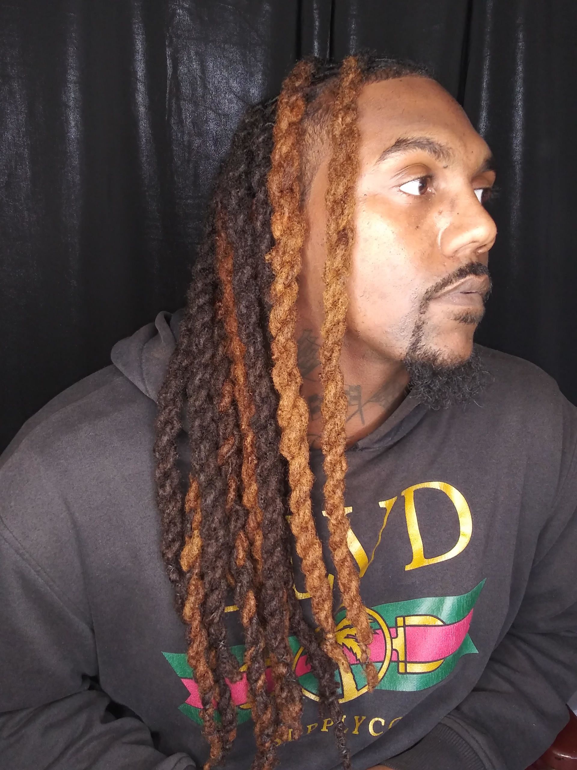 Person with long dreadlocks in a dark sweatshirt, looking to the right against a black background.