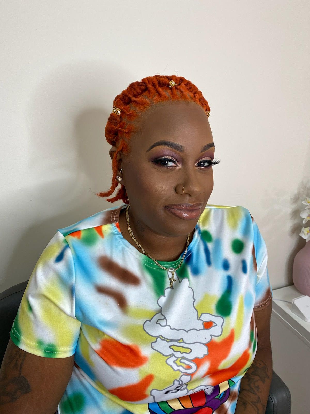 Person with orange hair wearing a colorful tie-dye shirt, seated against a plain wall.