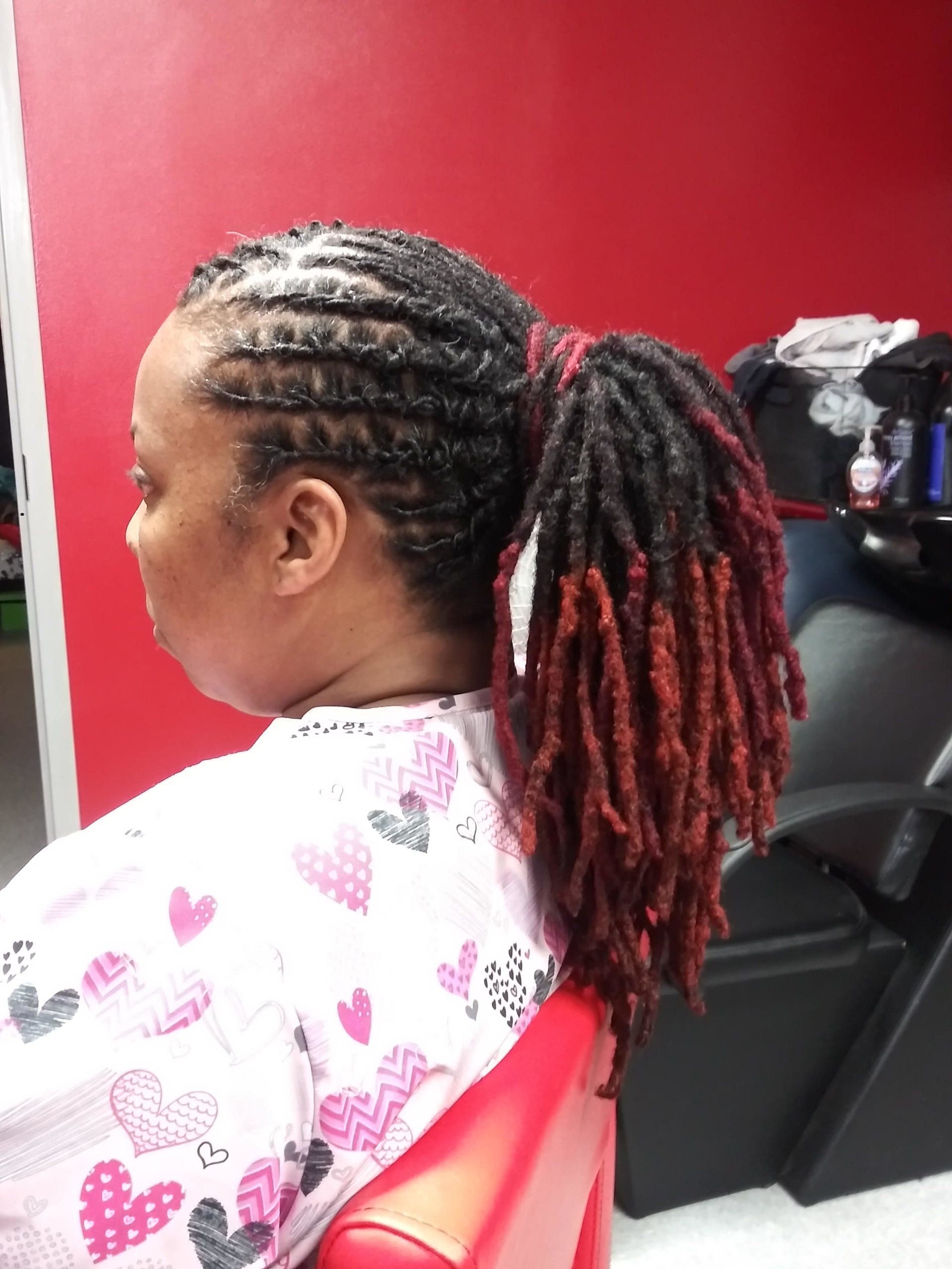 Woman with braided red-and-black hair seated in a salon chair against a red wall