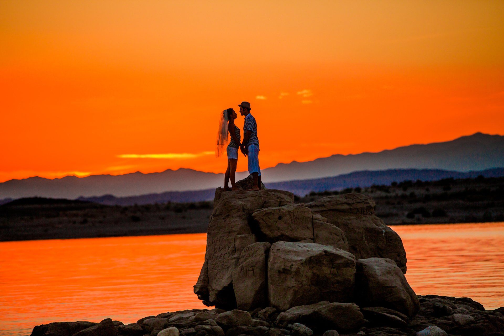 A man and a woman are standing on a rock overlooking a lake at sunset.