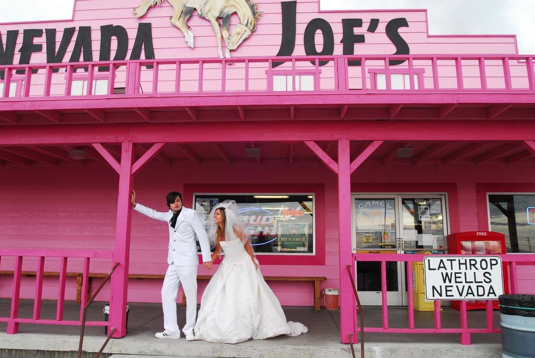 A bride and groom pose in front of a pink building called joe 's