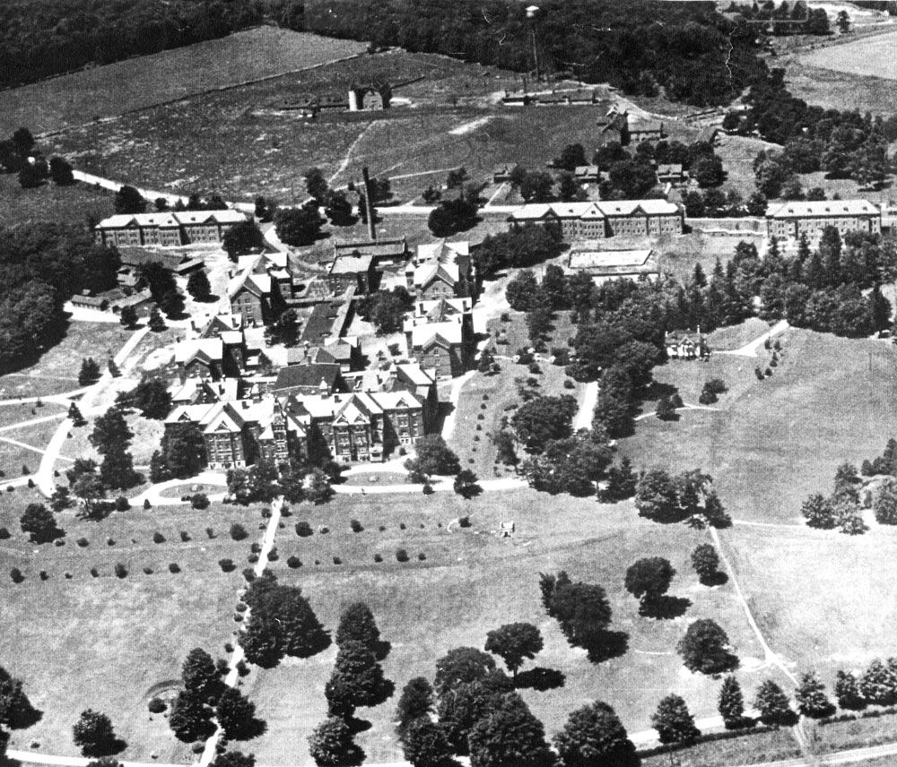 An aerial view of the Ontario Hospital School, Orillia as it appeared in the 1930s. Closest to the bottom of this image is the Administration Building, flanked on either side by Cottages K (left) and A (right). To the left of Cottage K is the newly constructed Hospital/Infirmary, which was used an admissions ward until the construction of the Isolation Ward in the mid-1950s. Behind the administration building is the Recreation Hall/Gymnasium and running back from this building is a long structure which contained facilities such as the institution kitchens and workshops. Between this structure and Memorial Avenue is the second institution Power House. On the left side of this long structure are Cottages L and M and on the right side are Cottages B and C. Running parallel to Memorial Avenue are three cottages constructed during S.J.W. Horne’s tenure as Superintendent. From left to right, these are Cottages O and D and the Nurses’ Residence. On the opposite side of the road is the institution farm. Three small farm houses face the road and beyond these, from left to right, are the cow barn, piggery and water tower, horse barn and blacksmith shop. To the right of the blacksmith shop is the institution cemetery. Image from the Regional Centre Records.) An aerial view of the Ontario Hospital School, Orillia as it appeared in the 1930s. Closest to the bottom of this image is the Administration Building, flanked on either side by Cottages K (left) and A (right). To the left of Cottage K is the newly constructed Hospital/Infirmary, which was used an admissions ward until the construction of the Isolation Ward in the mid-1950s. Behind the administration building is the Recreation Hall/Gymnasium and running back from this building is a long structure which contained facilities such as the institution kitchens and workshops. Between this structure and Memorial Avenue is the second institution Power House. On the left side of this long structure are Cottages L and M and on the right side are Cottages B and C. Running parallel to Memorial Avenue are three cottages constructed during S.J.W. Horne’s tenure as Superintendent. From left to right, these are Cottages O and D and the Nurses’ Residence. On the opposite side of the road is the institution farm. Three small farm houses face the road and beyond these, from left to right, are the cow barn, piggery and water tower, horse barn and blacksmith shop. To the right of the blacksmith shop is the institution cemetery. Image from the Regional Centre Records.)