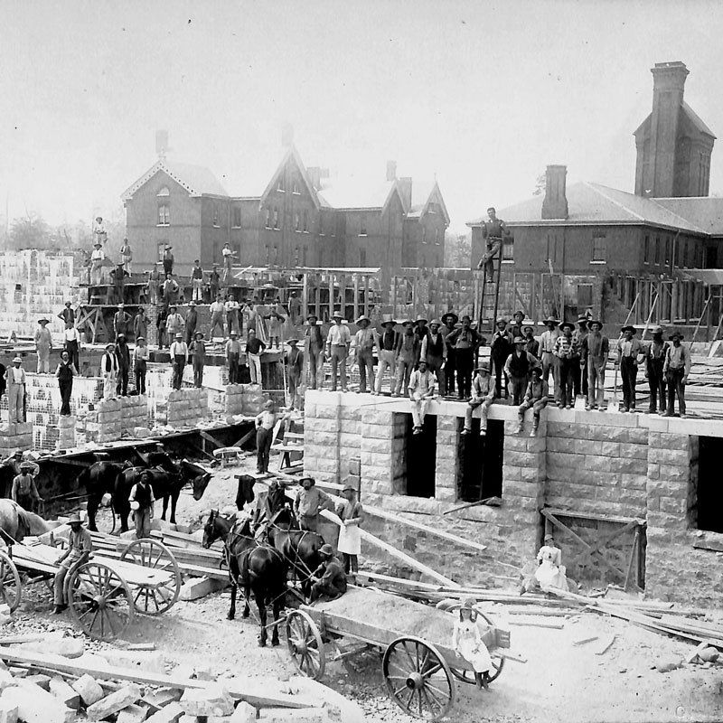 A photograph showing the construction of Cottages A and K and the Administration Building. Workers stand and sit on the building’s stone foundations and three teams of horses and two wagons are standing on the ground below. Behind the foundations, Cottage L, a large three storey brick building, is visible, as is the institution’s original powerhouse and chimney. Image from the Orillia Public Library. A photograph showing the construction of Cottages A and K and the Administration Building. Workers stand and sit on the building’s stone foundations and three teams of horses and two wagons are standing on the ground below. Behind the foundations, Cottage L, a large three storey brick building, is visible, as is the institution’s original powerhouse and chimney. Image from the Orillia Public Library.