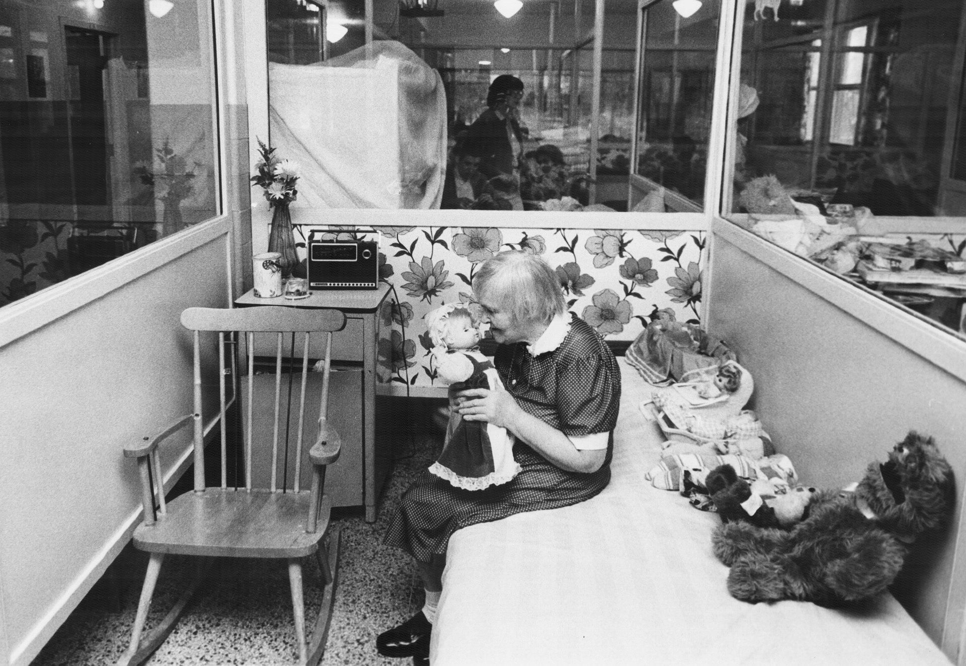 Living conditions at the Huronia Regional Centre in 1982. The image shows a small cubicle with walls mostly made out of clear glass. One side of the cubicle is taken up by a narrow bed, on which there is a row of dolls and stuffed toys. Next to this bed is a wooden rocking chair and a small night table with items on top of it, including a vase of flowers and a radio. An elderly woman is sitting on the bed, holding a doll. Given her age, she was likely institutionalized as a child in the early 20th century and spent her entire life in the facility. Image from the Toronto Star Archives. Living conditions at the Huronia Regional Centre in 1982. The image shows a small cubicle with walls mostly made out of clear glass. One side of the cubicle is taken up by a narrow bed, on which there is a row of dolls and stuffed toys. Next to this bed is a wooden rocking chair and a small night table with items on top of it, including a vase of flowers and a radio. An elderly woman is sitting on the bed, holding a doll. Given her age, she was likely institutionalized as a child in the early 20th century and spent her entire life in the facility. Image from the Toronto Star Archives.