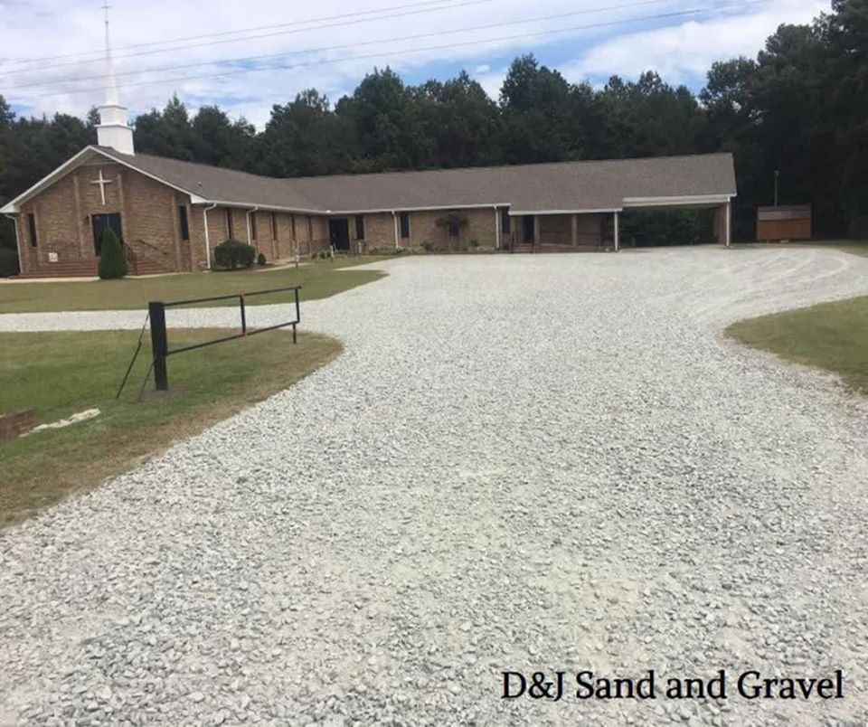 Gravel driveway leading to a brick church with a steeple. Grass and trees surround the building.