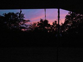 Silhouetten van bomen tegen een roze en paarse zonsondergang, gezien vanaf een schaduwrijke veranda.