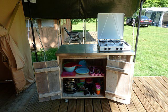 Buitenkeuken met fornuis op een houten aanrecht en open kastplanken met kommen en kookgerei.
