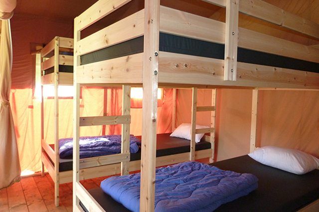 Houten stapelbedden in een slaapzaal met blauwe matrassen en warme gordijnen voor de ramen.
