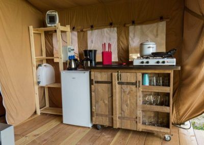Een kleine keukenopstelling met houten kastjes, een minikoelkast, een waterkoker, een kookplaat en planken in een tent.