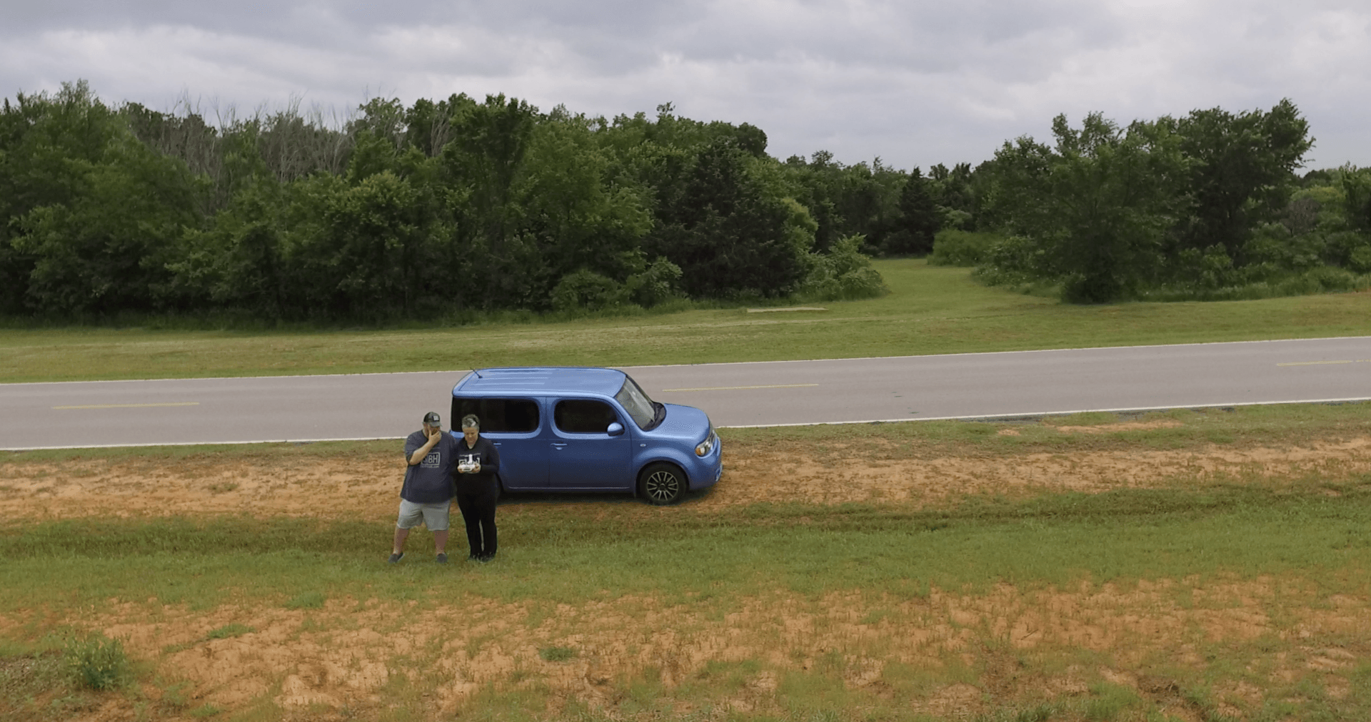 man and woman in feild next to parked blue cube car flying drone, (mitch park, edmond oklahoma)
