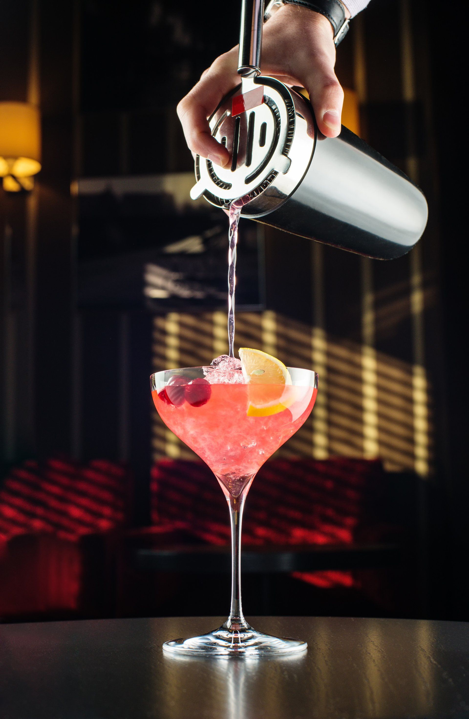 Bartender pouring a pink cocktail into a stemmed glass, garnished with a lemon and berries, in a dim bar.