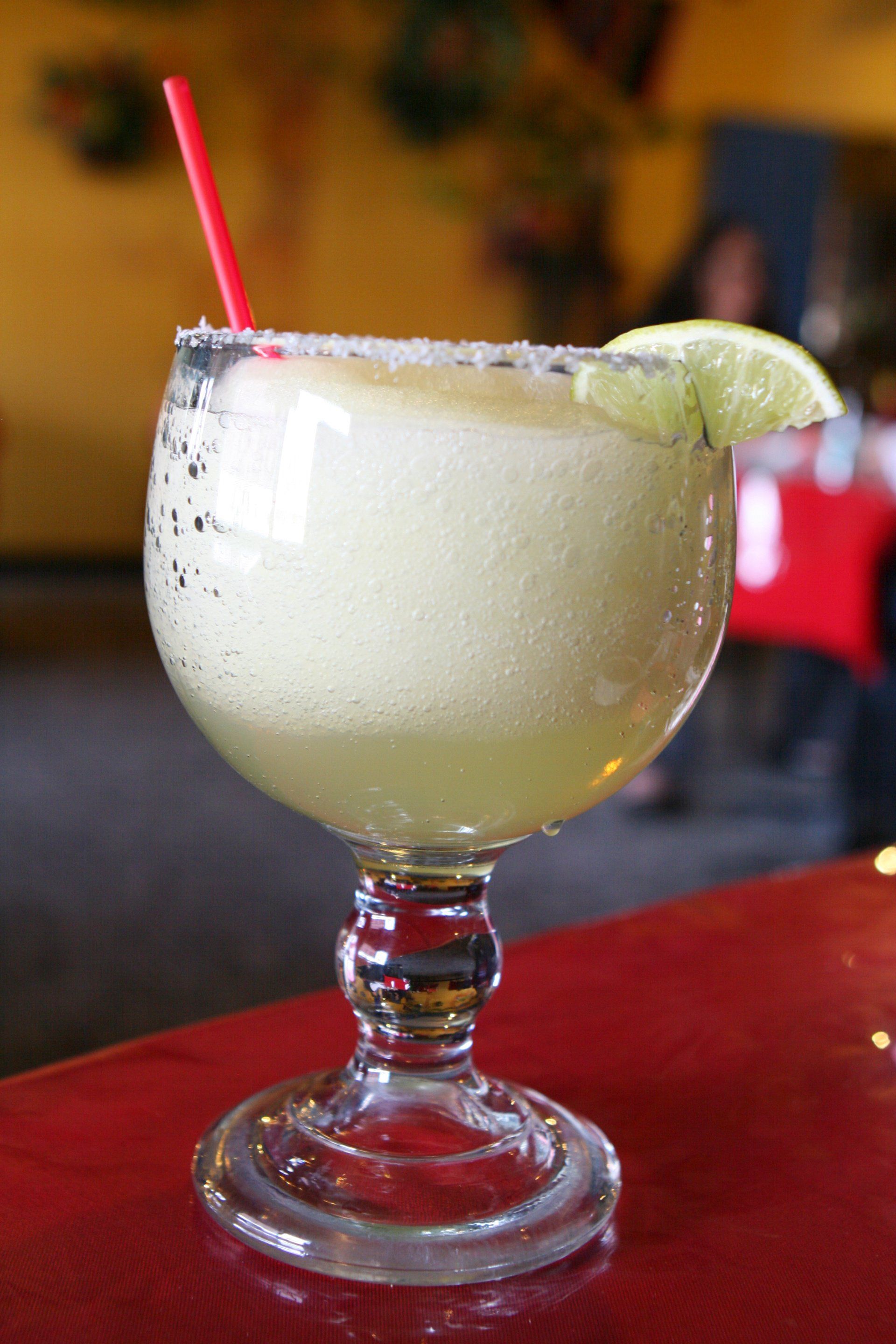 Frozen margarita in a stemmed glass with a lime slice and red straw, on a red table.