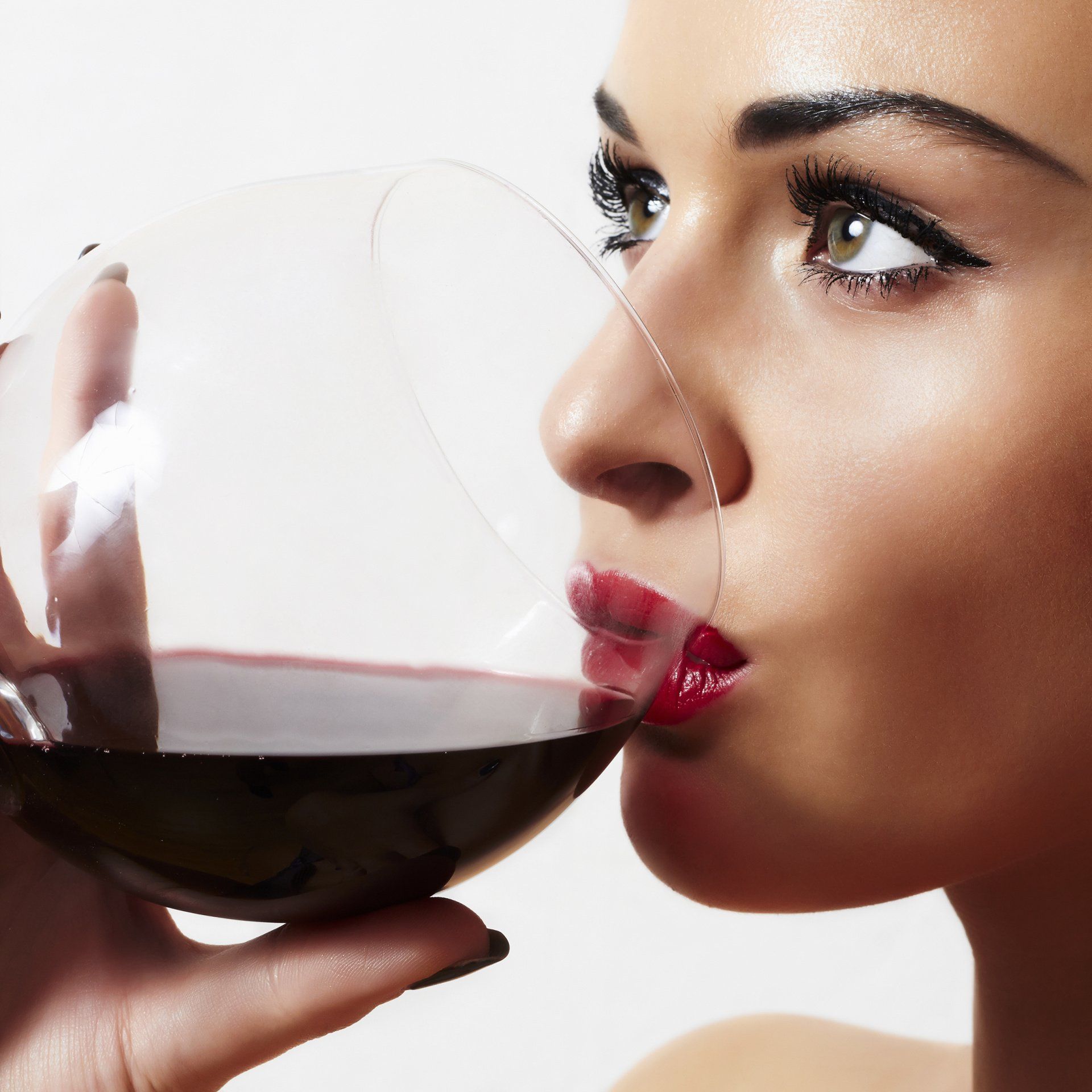 Woman sipping red wine from a large glass, against a white background.
