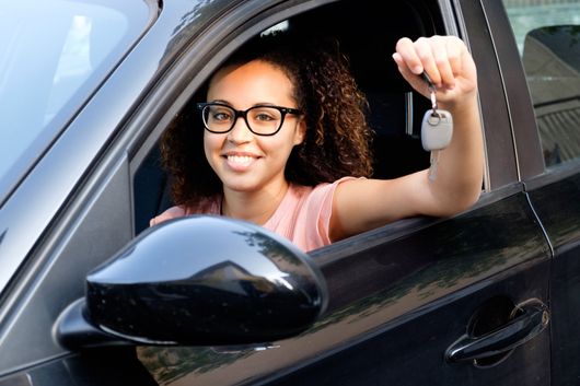 A smiling person sitting in the driver's seat of a black car, holding up a set of car keys. A smiling person sitting in the driver's seat of a black car, holding up a set of car keys.