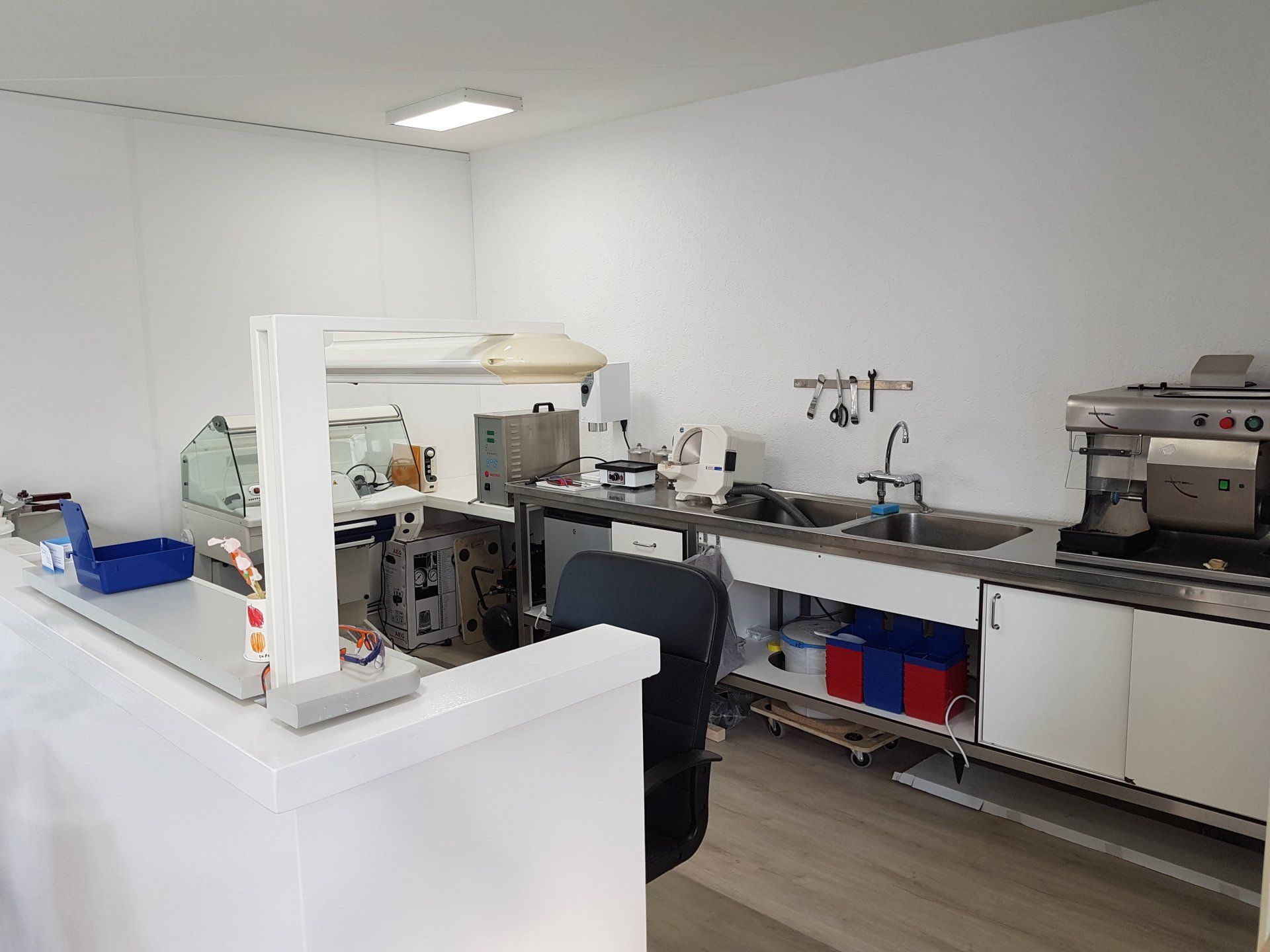 Een klinische laboratoriumwerkplek met een wit bureau, een wastafel, medische apparatuur en een zwarte bureaustoel in een schone, witte ruimte.