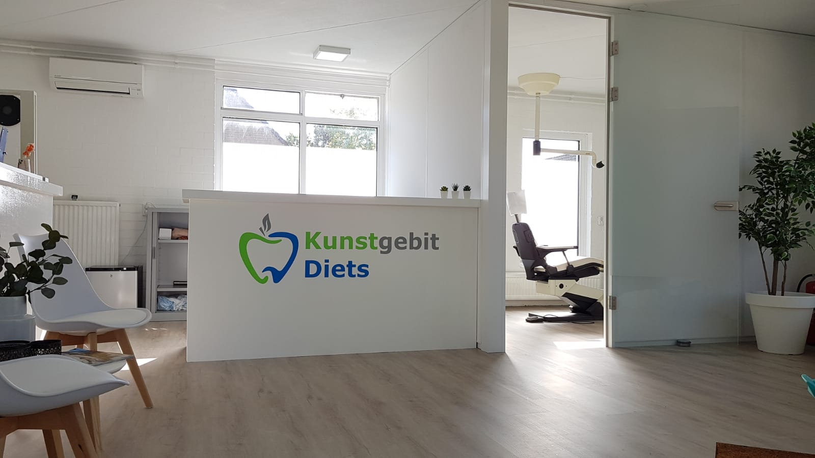 De ontvangsthal van een tandartspraktijk met een logo op de balie en zicht op een behandelkamer met een stoel.