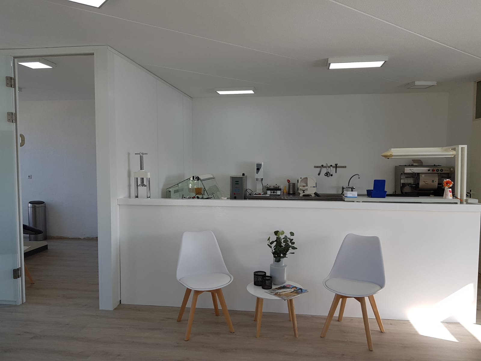 Een moderne, minimalistische wachtruimte met twee witte stoelen, een kleine tafel en een servicebalie in een lichte ruimte.