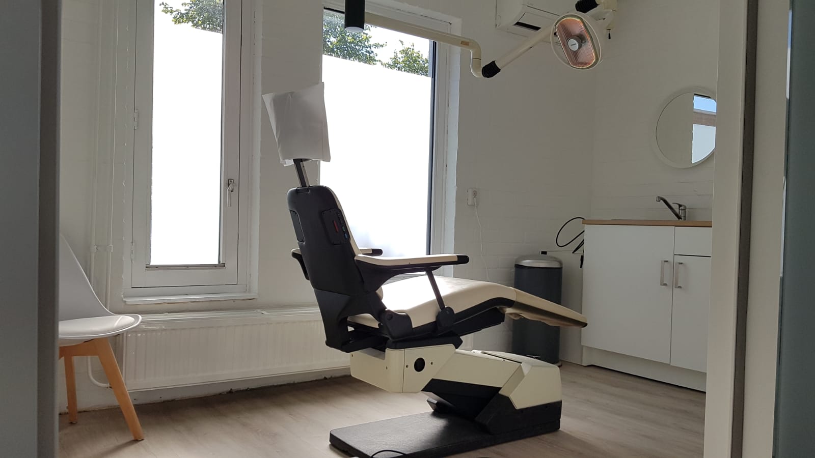 Een behandelkamer is voorzien van een professionele medische stoel in het midden tussen twee ramen, met een wastafel en een badmeubel rechts ervan.
