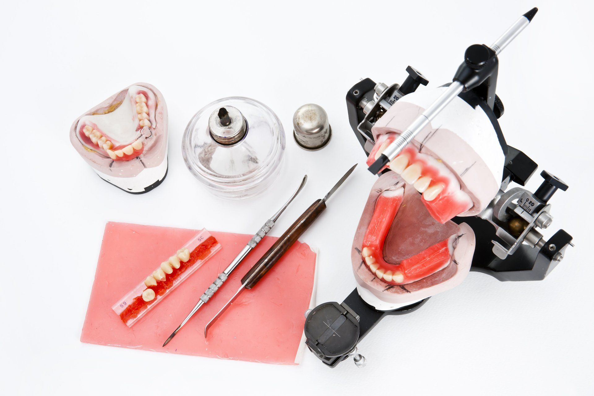 Een tandheelkundige articulator met gebitsprothese, een model, metalen instrumenten en wasmateriaal op een witte achtergrond.