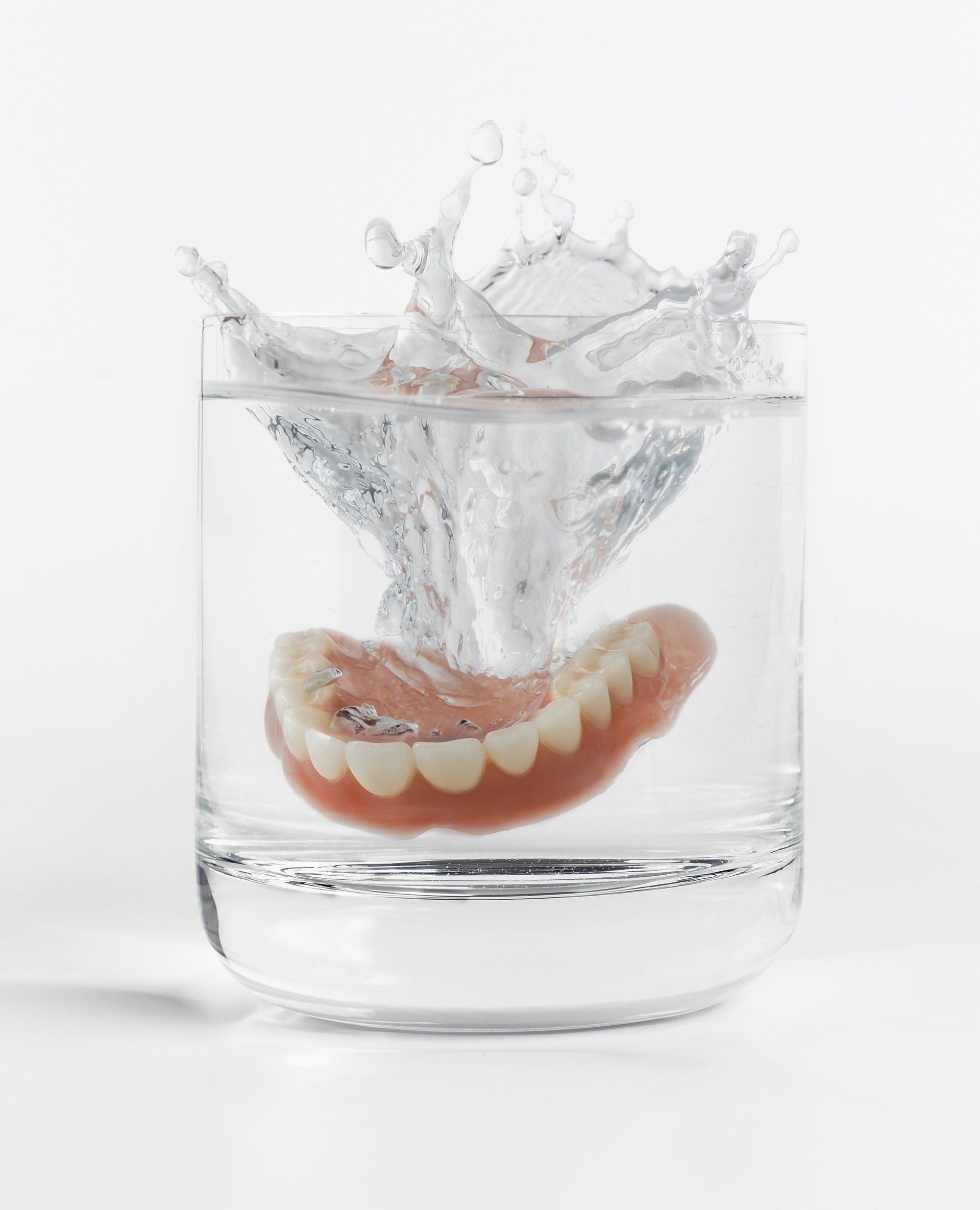 Een kunstgebit ondergedompeld in een helder glas water, waardoor er spetters aan het oppervlak ontstaan ​​tegen een witte achtergrond.