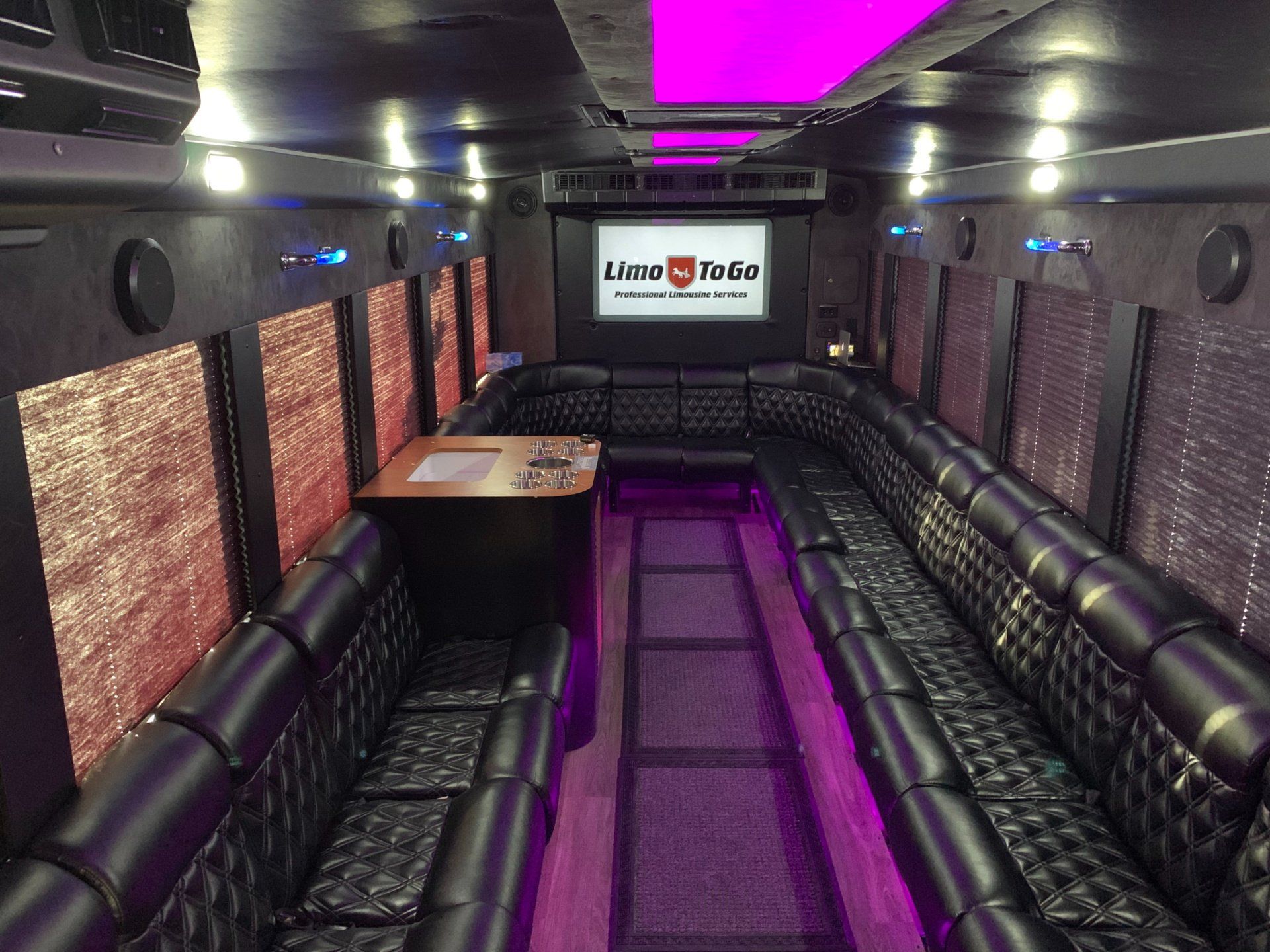 white International 26 passenger party bus interior with black tufted plush seating and purple LED lights and Limo To Go logo displayed on back big screen TV