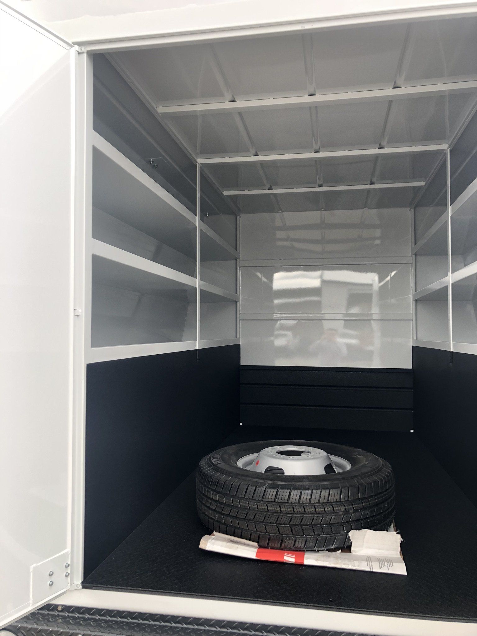 Inside of an empty cargo van, white walls, shelving, black floor, spare tire.