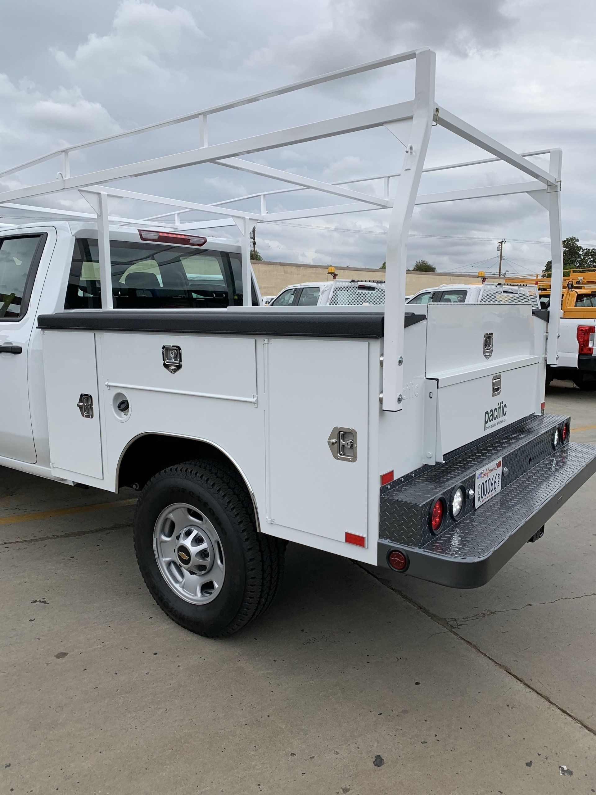 White work truck with ladder rack, toolboxes, and diamond-plate bumper parked on concrete.