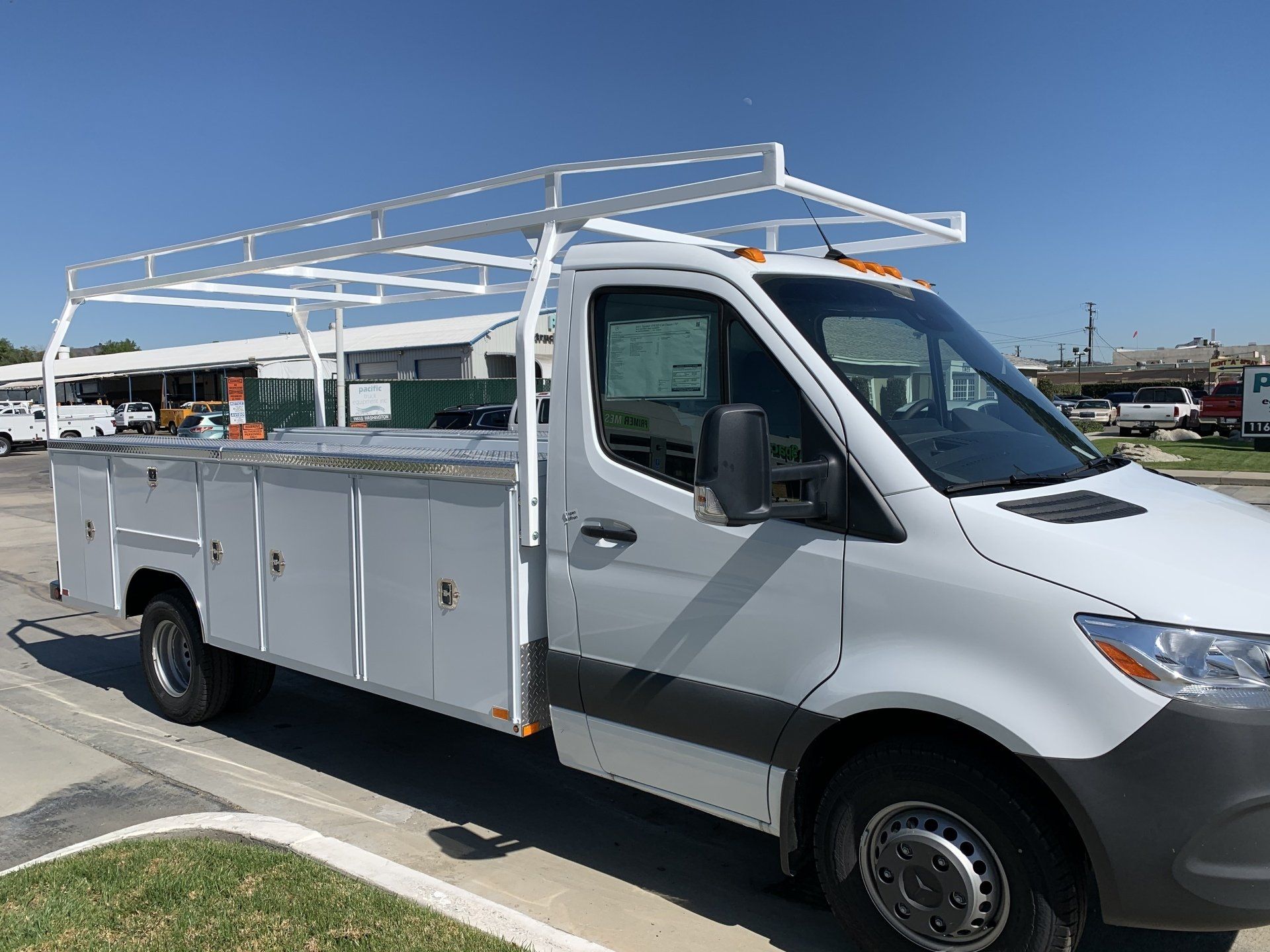 White work van with ladder rack, side storage compartments, parked outdoors.