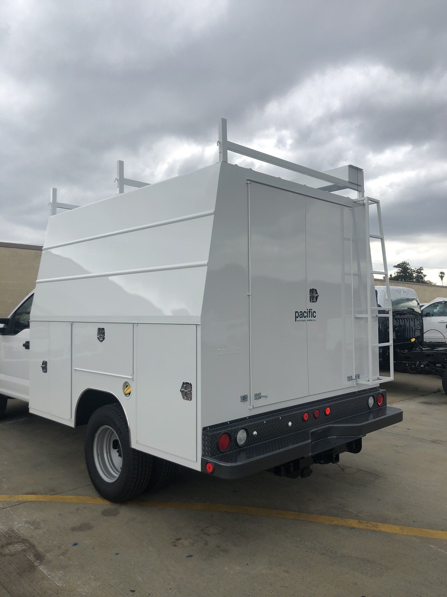 White utility truck with enclosed bed, ladder rack, and rear door open on an overcast day.