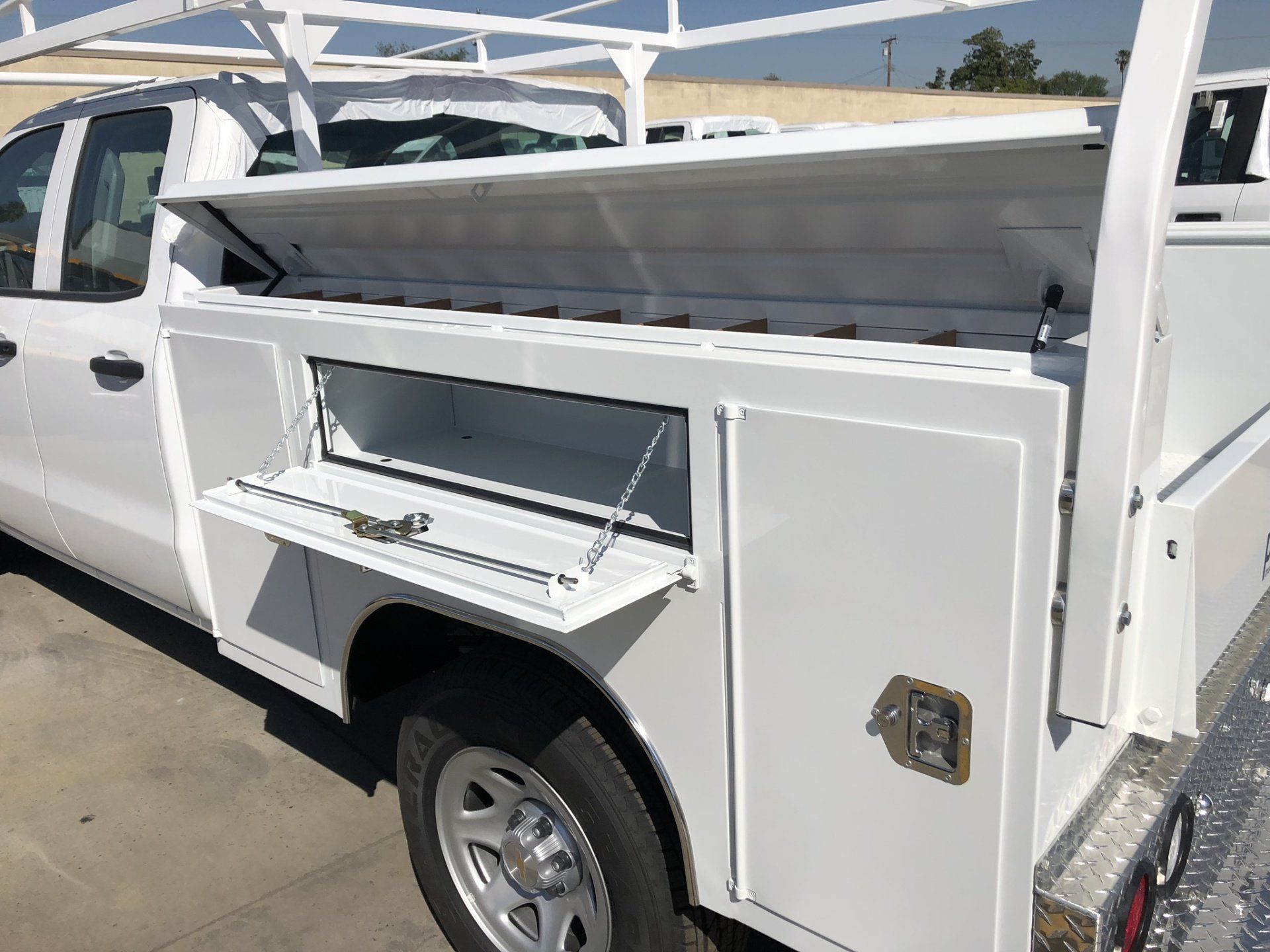 White utility truck with open storage compartments and ladder rack, parked outdoors.