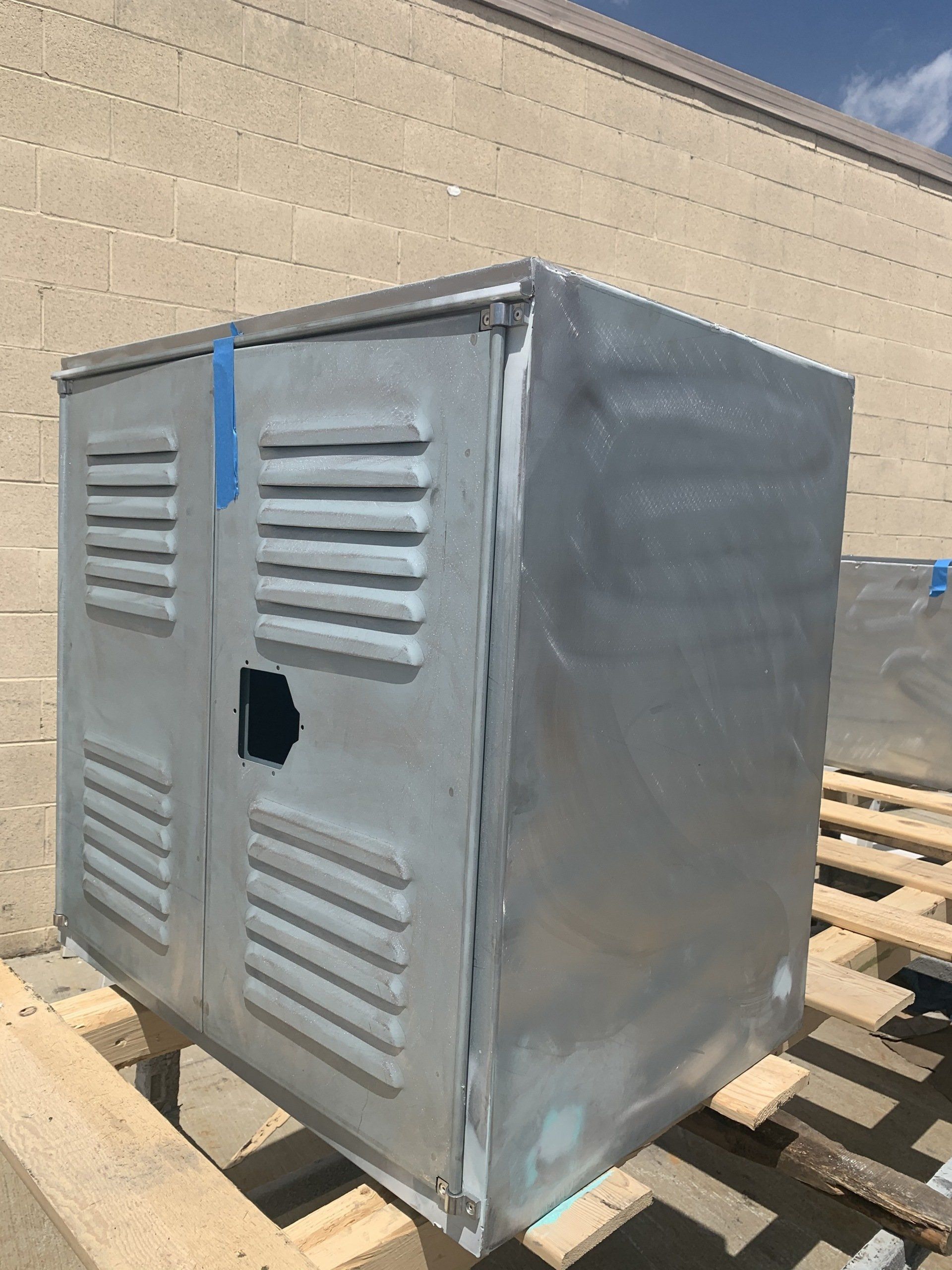 Metal utility box with ventilation slots, taped, on a wooden pallet near a beige brick wall.
