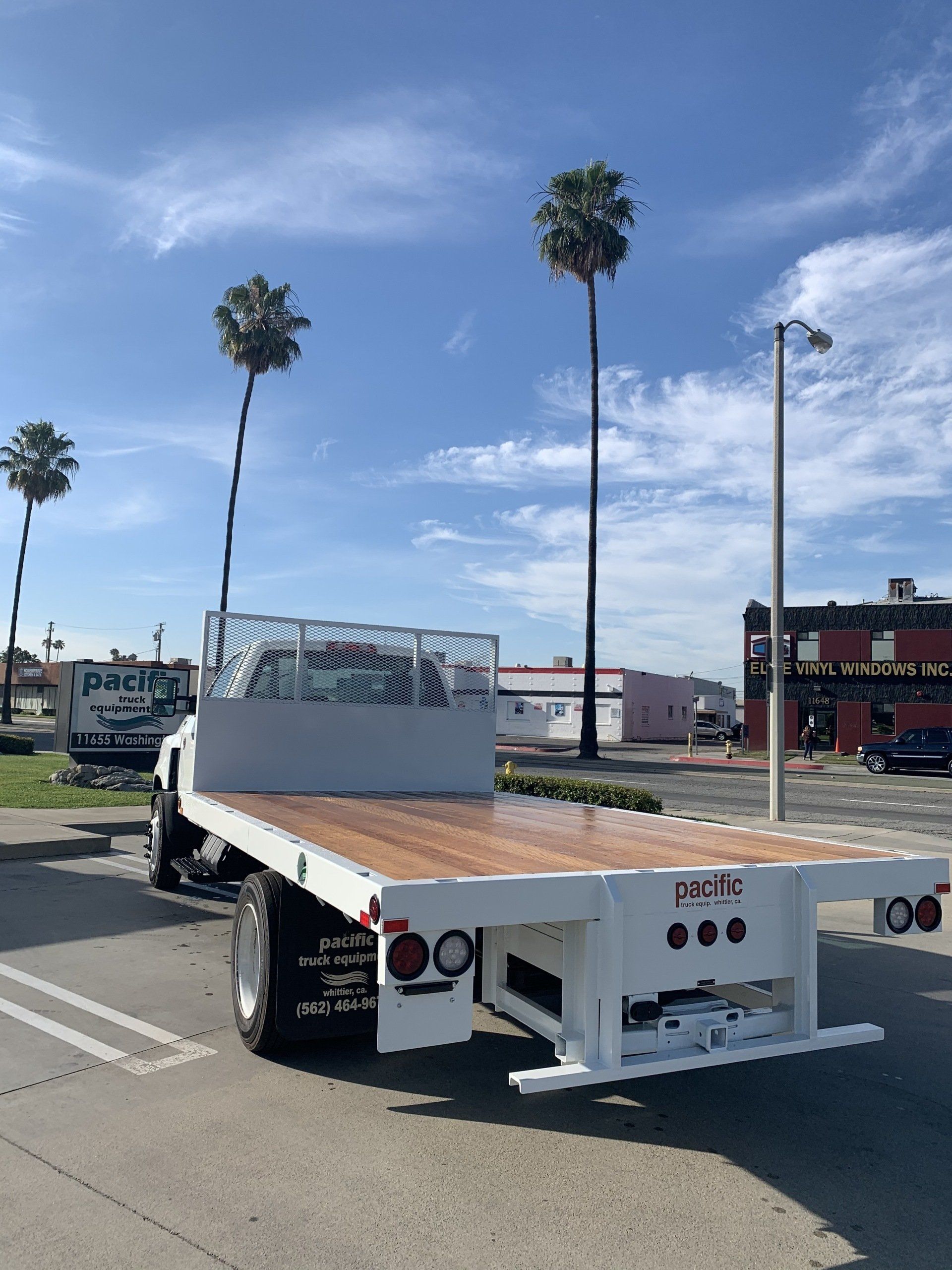 White flatbed truck on asphalt, with a wooden deck, under a blue sky with palm trees.