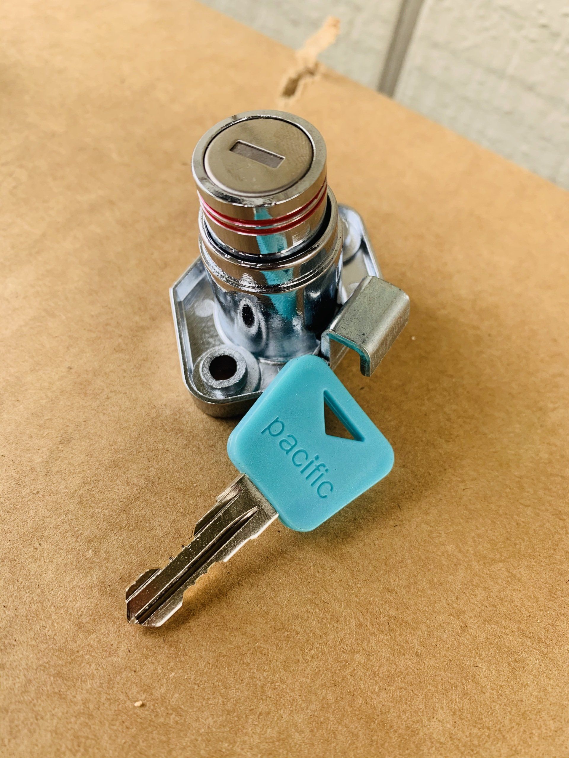 A chrome cabinet lock with a blue Pacific key on a cardboard surface.