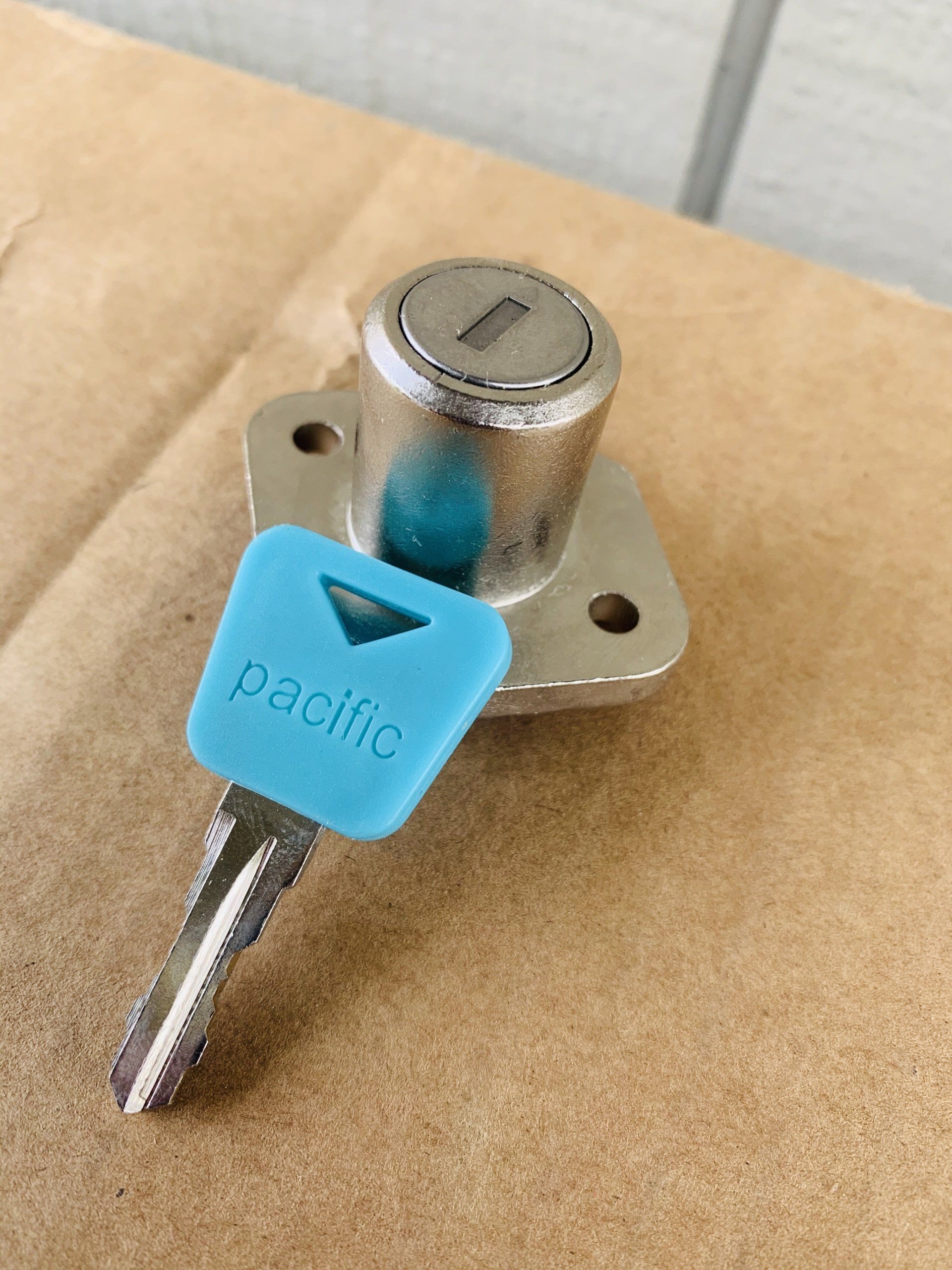 A silver cabinet lock with a blue Pacific key on a brown surface. The lock has a triangular base.