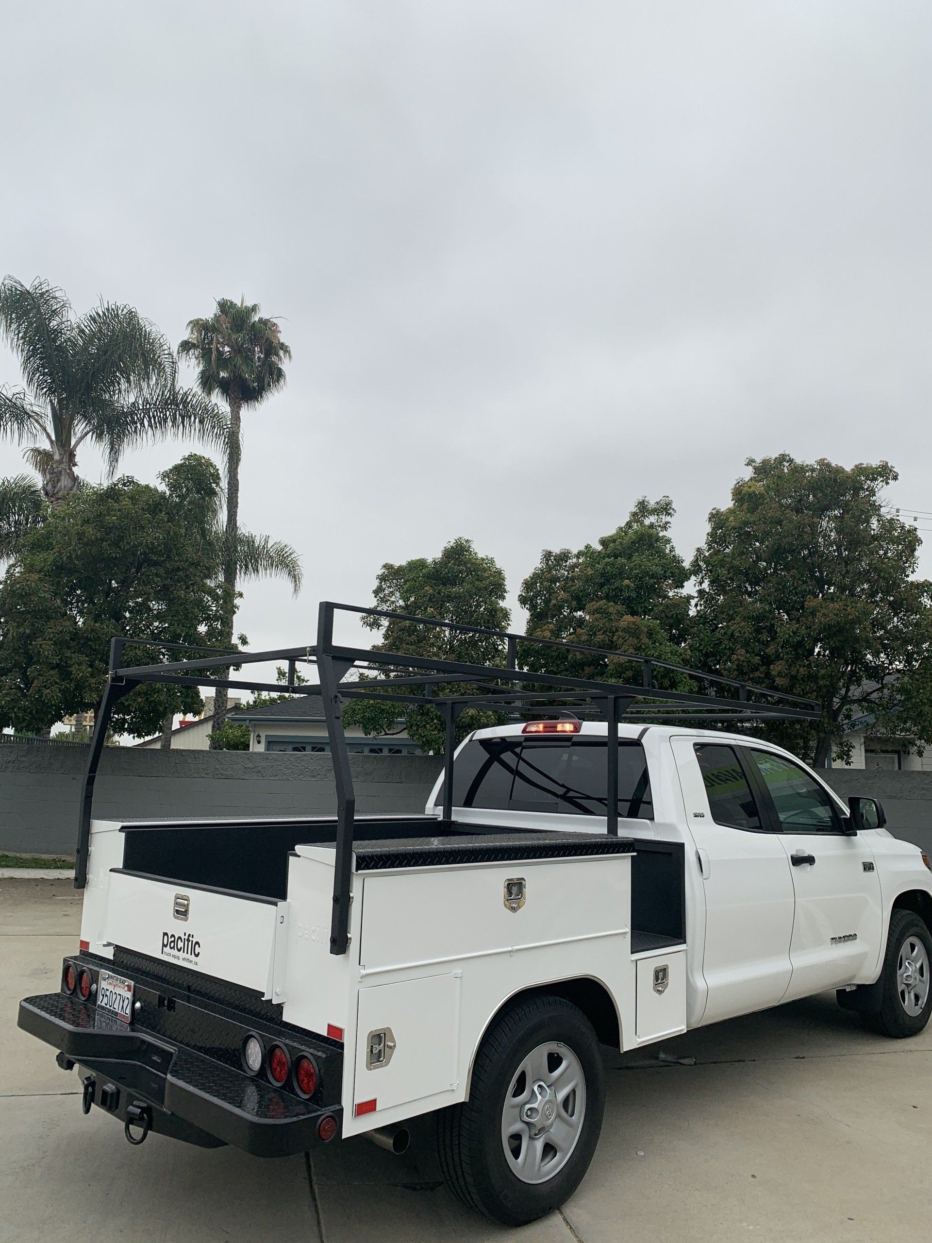 White work truck with black ladder rack and storage, parked outdoors on a cloudy day.
