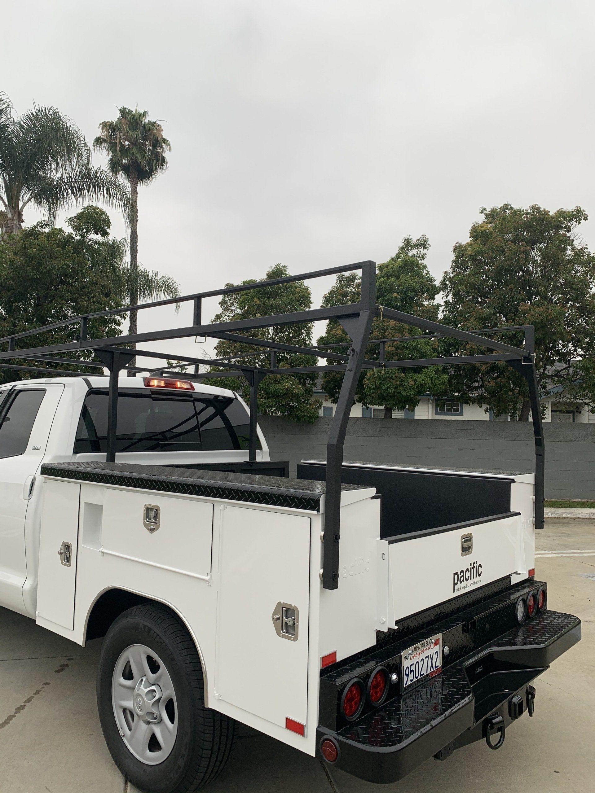White truck with black utility bed and ladder rack, parked outdoors on a cloudy day.