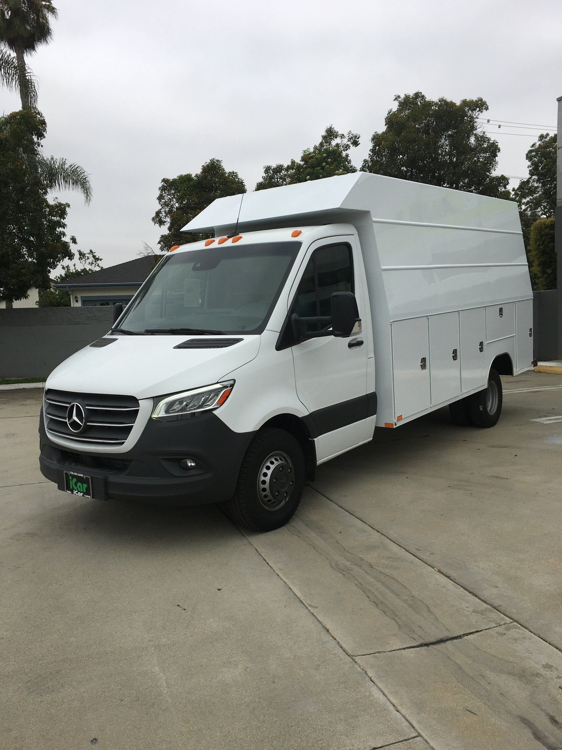White Mercedes-Benz Sprinter work van with custom utility body, parked on concrete. Cloudy day.