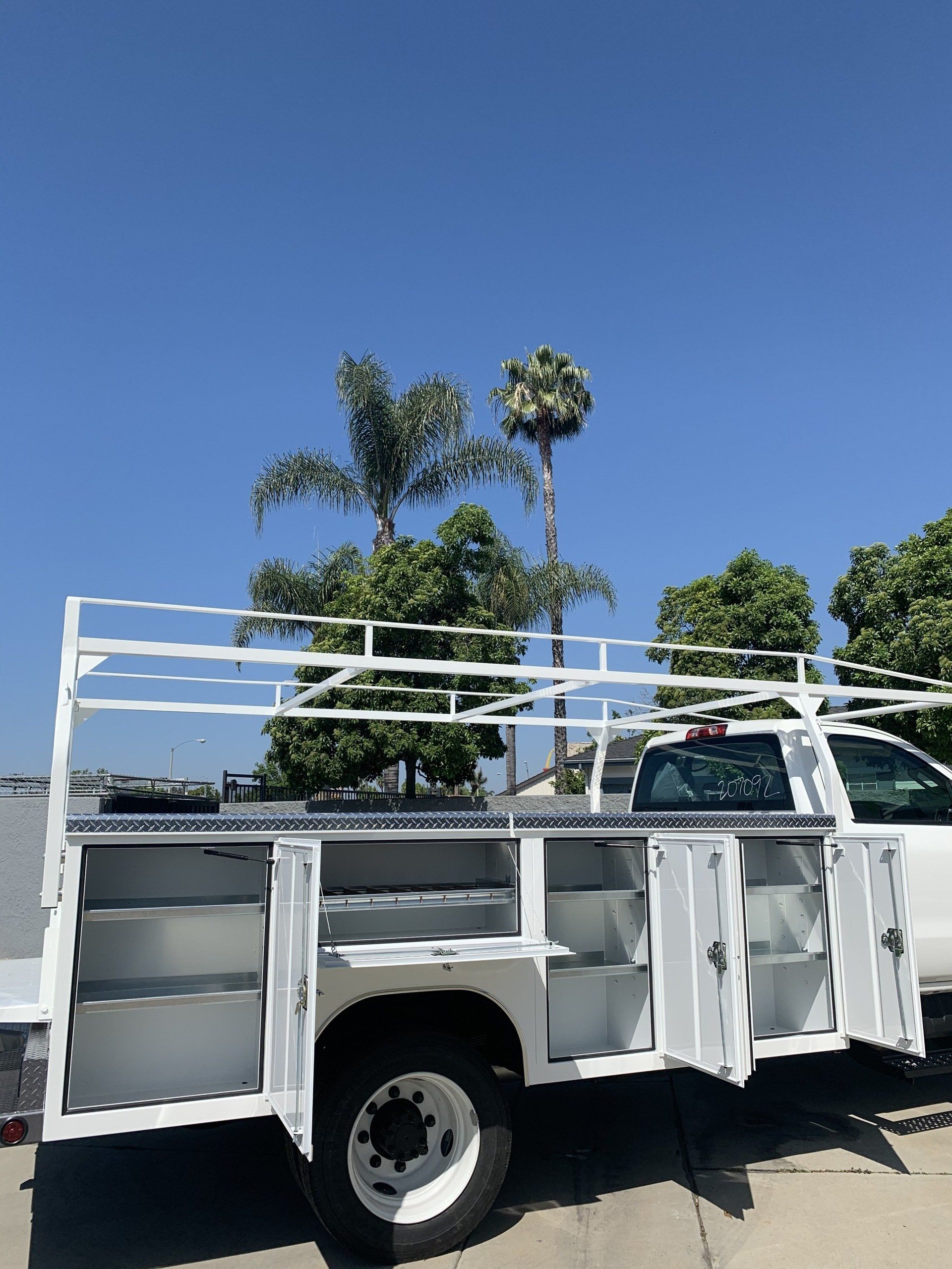 White work truck with open compartments and ladder rack, under a blue sky with palm trees.