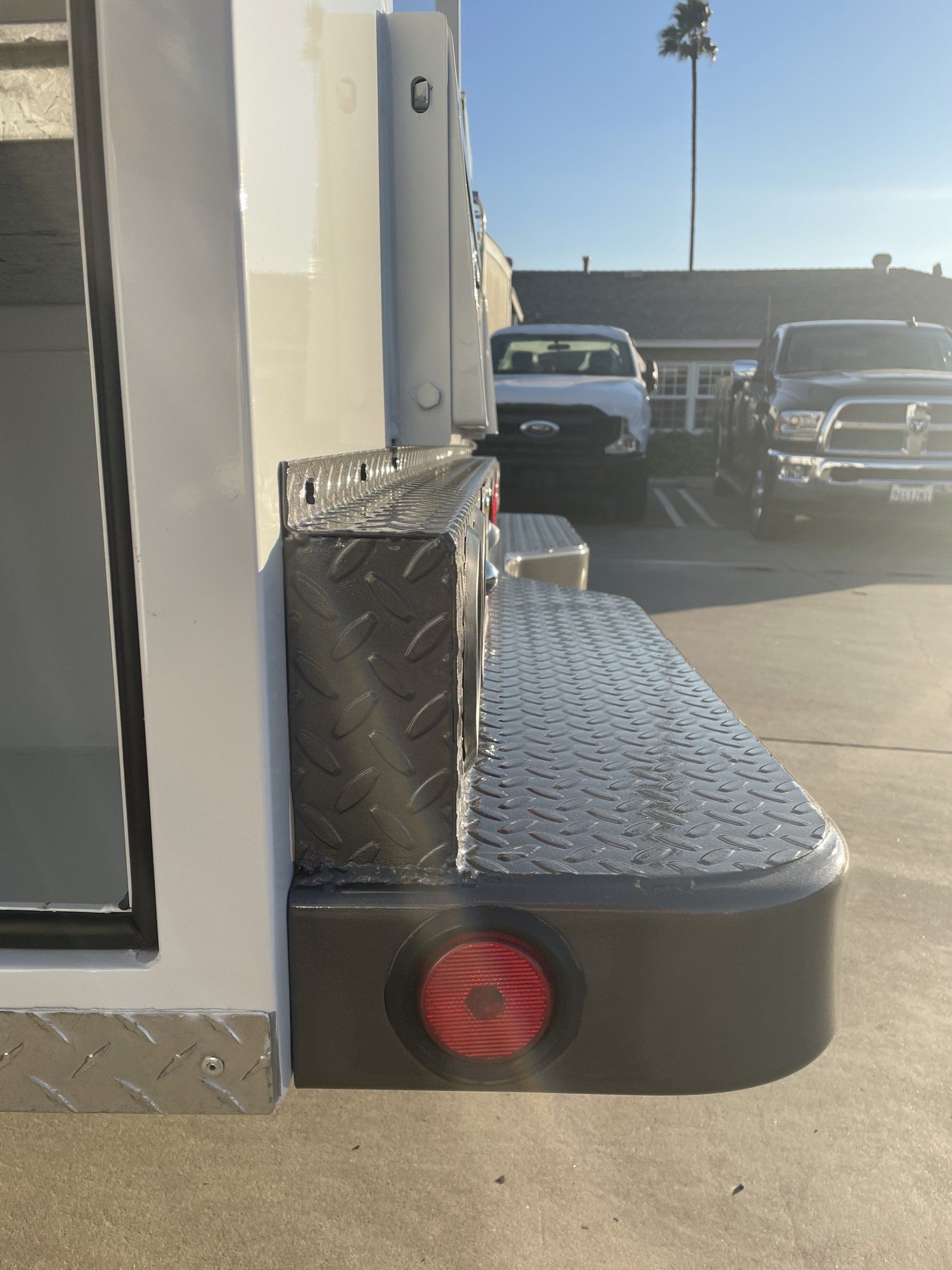 Diamond-plated step and storage box on the back of a white truck, with a red tail light.