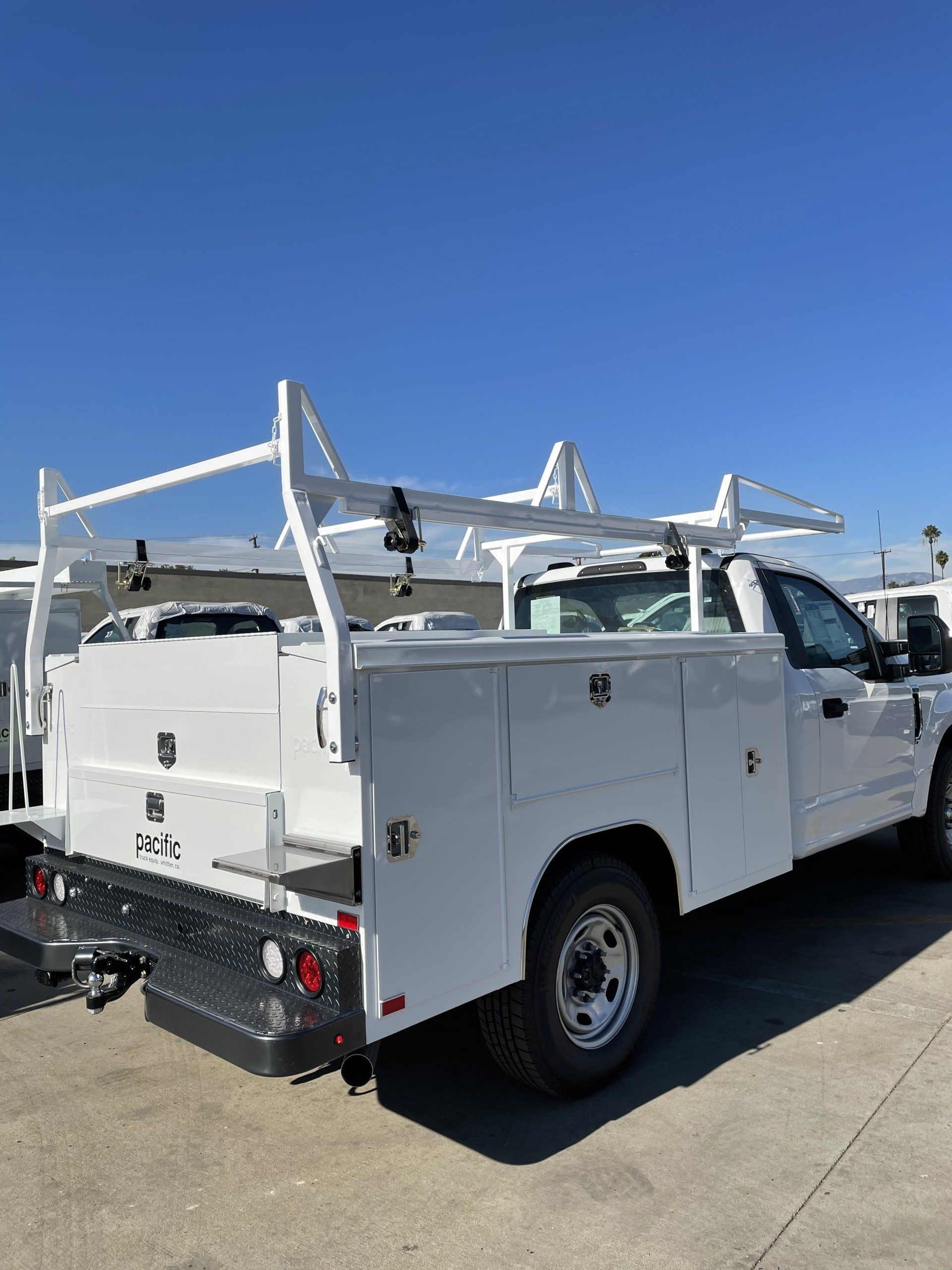 White work truck with ladder rack, toolboxes, and flatbed parked under a clear blue sky.