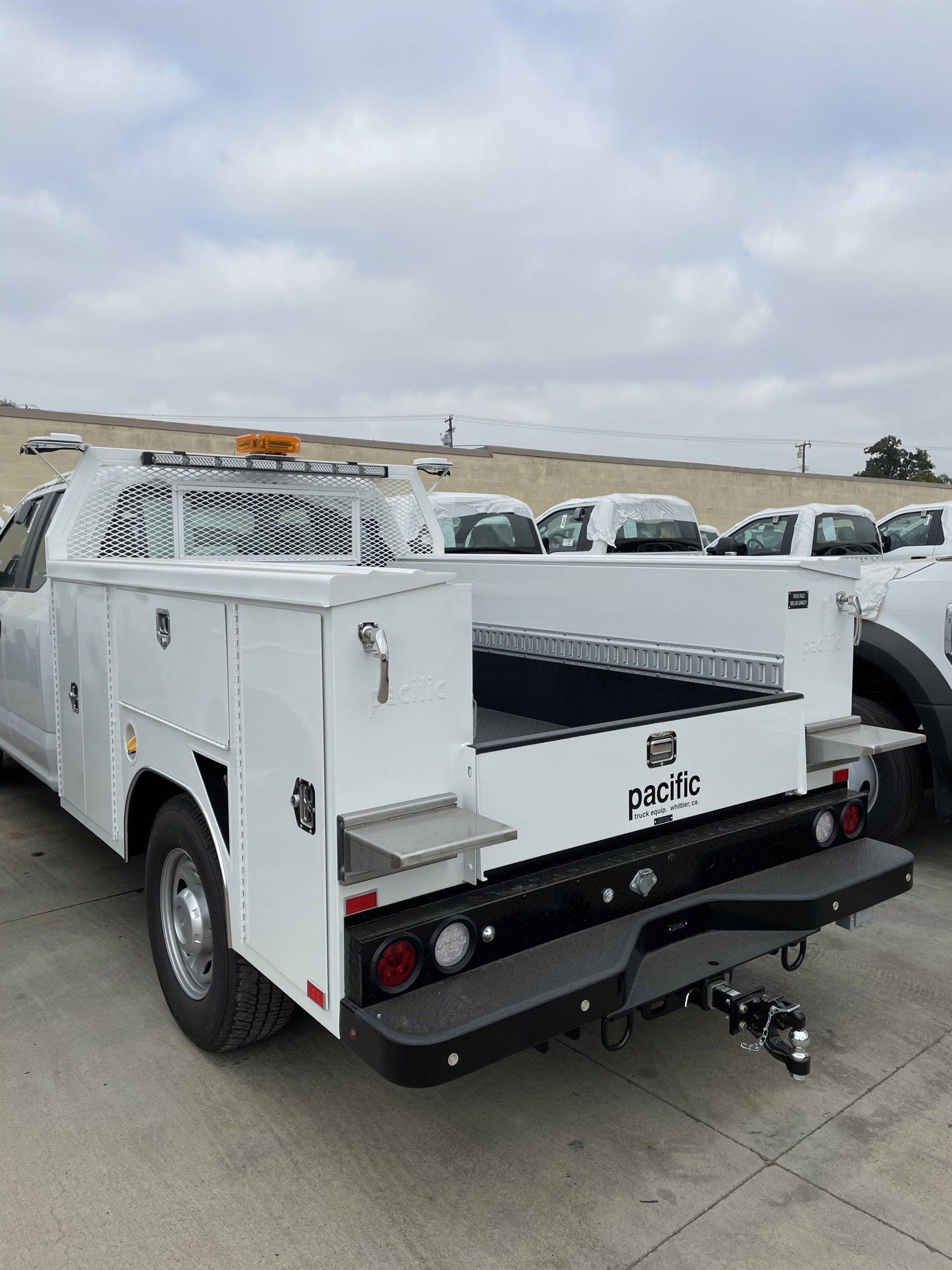 White utility truck with toolboxes and towing hitch, parked outdoors under a cloudy sky.