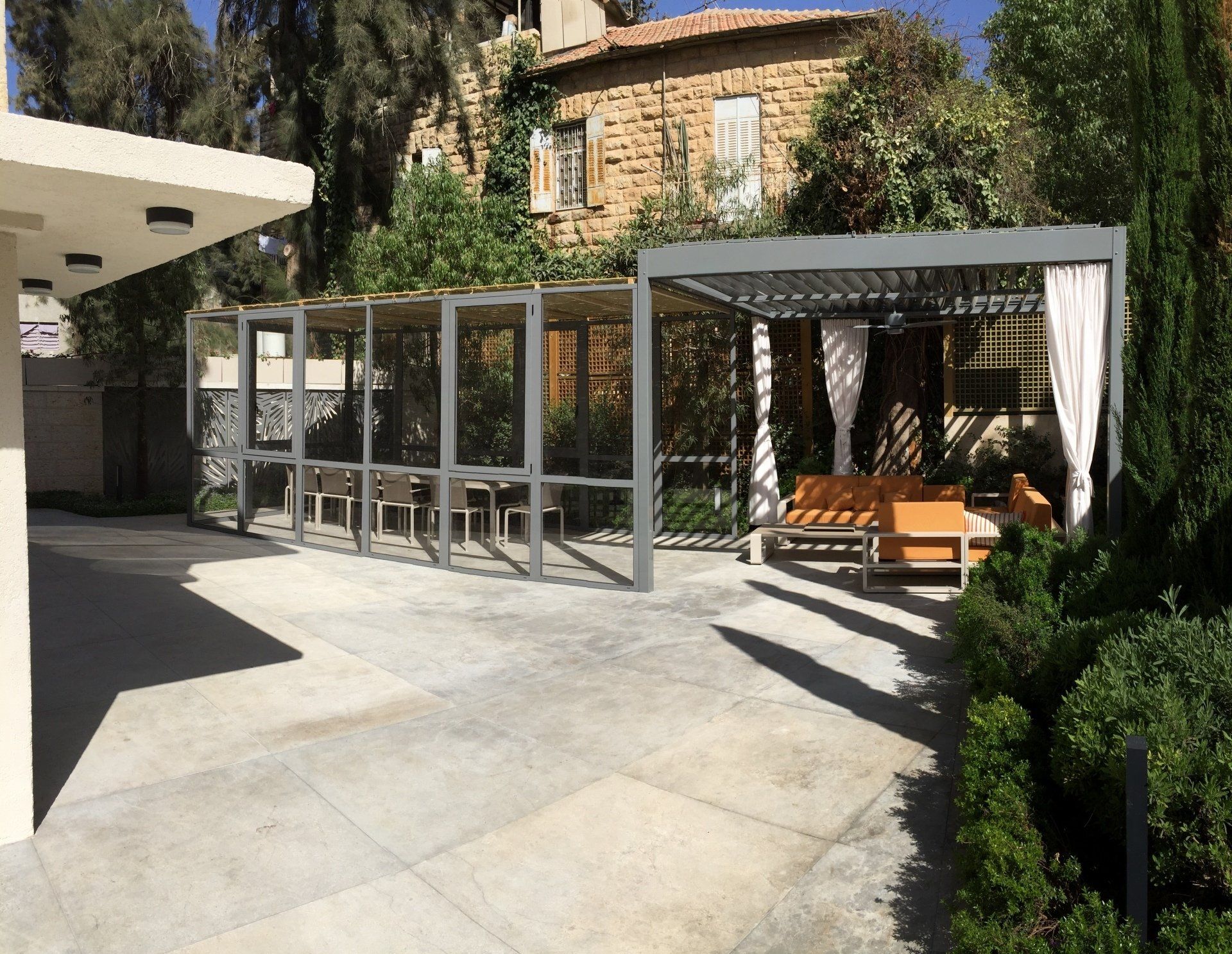 Modern patio with gray pergola, outdoor seating, and white curtains beside a stone building