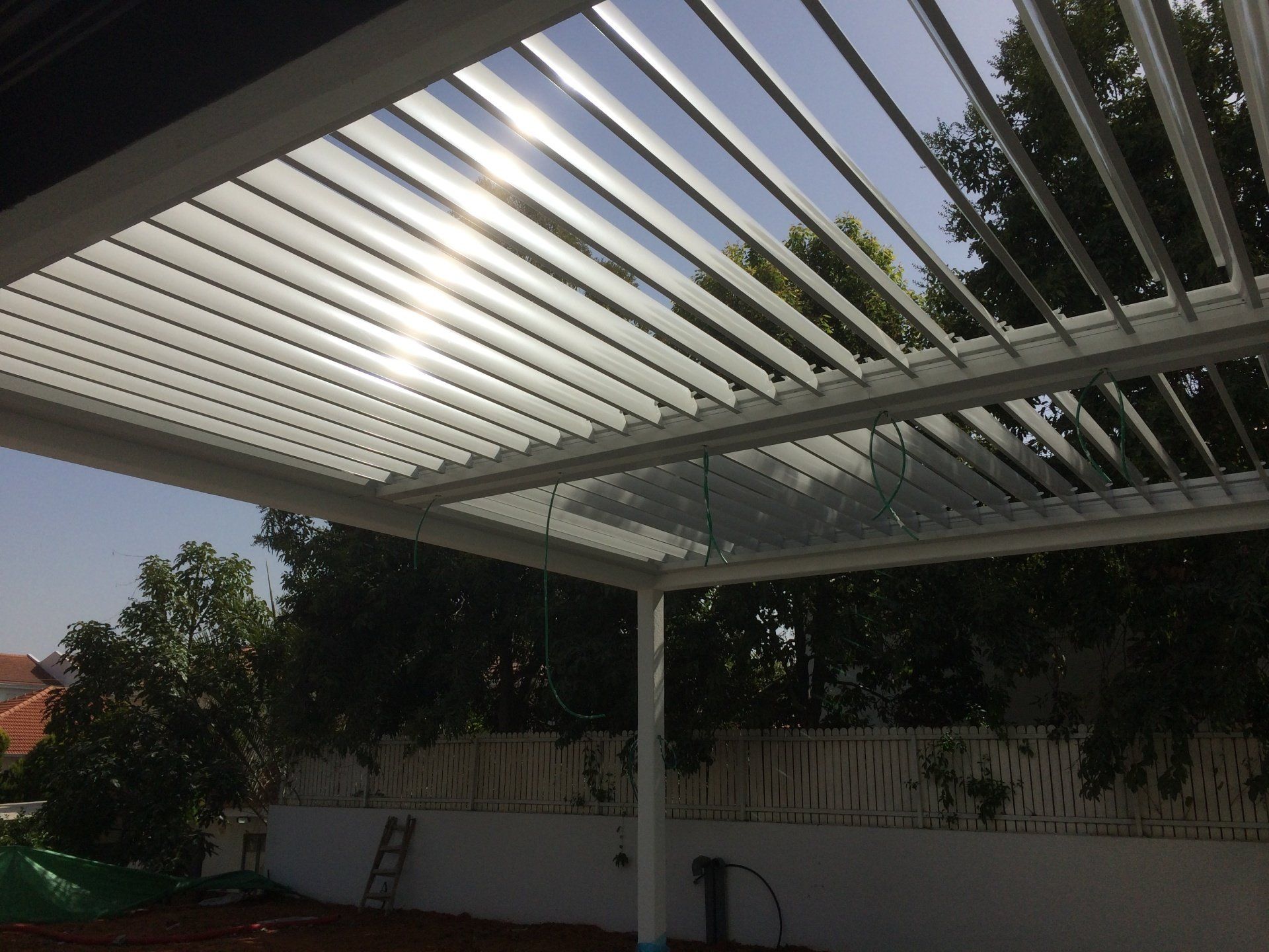 White slatted patio pergola over a backyard patio with trees and a ladder against a wall