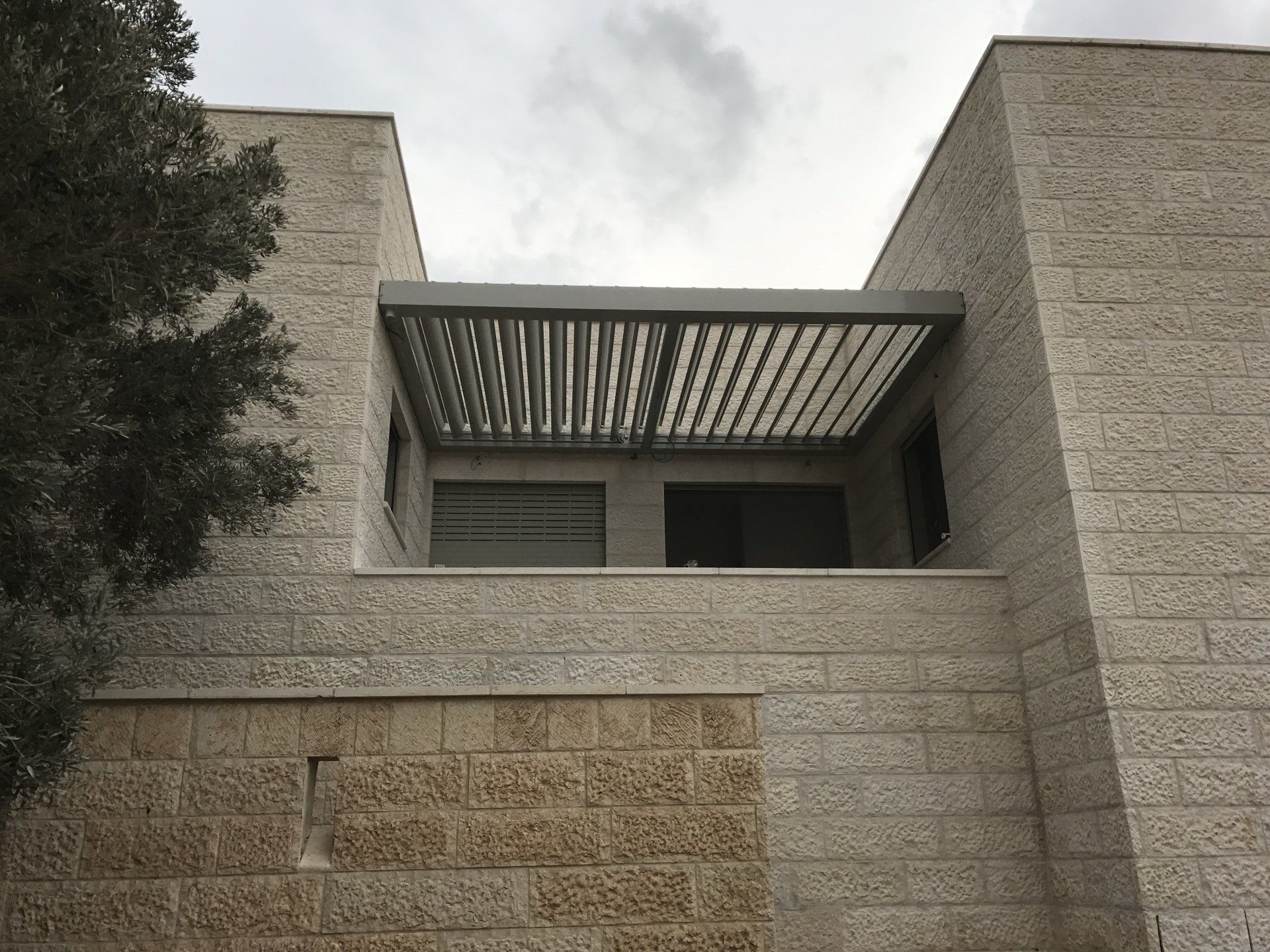 Stone building facade with a small balcony and slatted pergola under an overcast sky